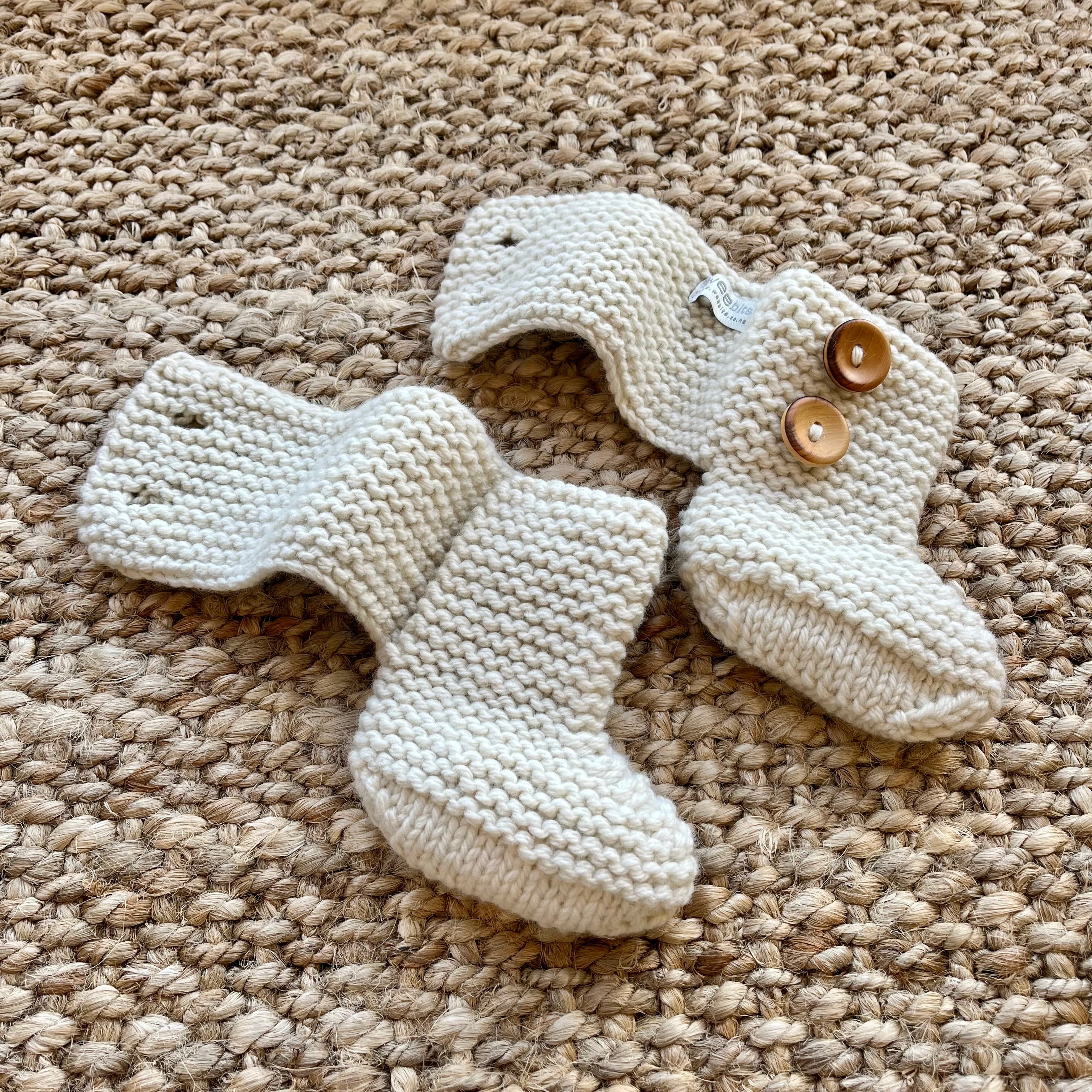 A pair of cream-colored hand-knit baby booties with wooden buttons, laid flat on a textured beige jute rug.
