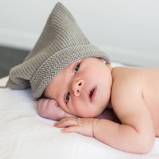 A baby lying on a white blanket wearing a gray knit pointed hat with a pom-pom.
