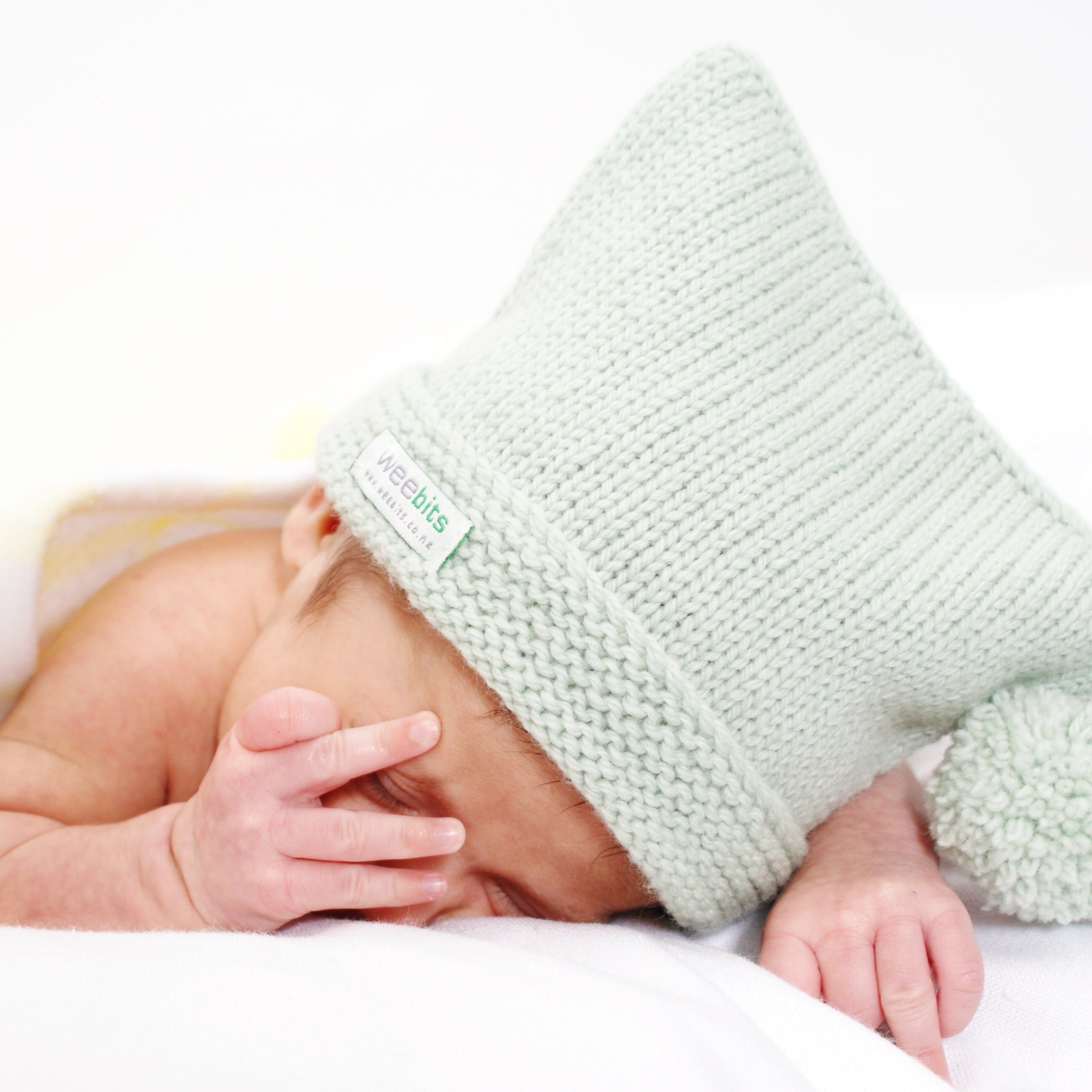 A newborn baby sleeping peacefully under a white blanket, wearing a pale sage green knitted beanie with a pom-pom. The beanie features a small white fabric label on the side.