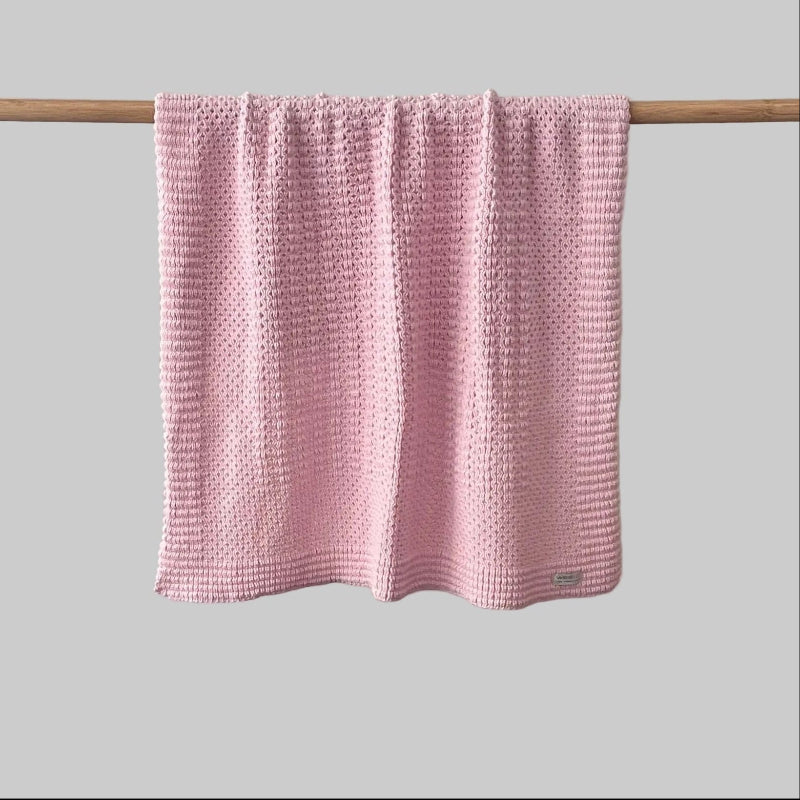 Textured blossom pink bassinet blanket hanging on a wooden rod against a grey background