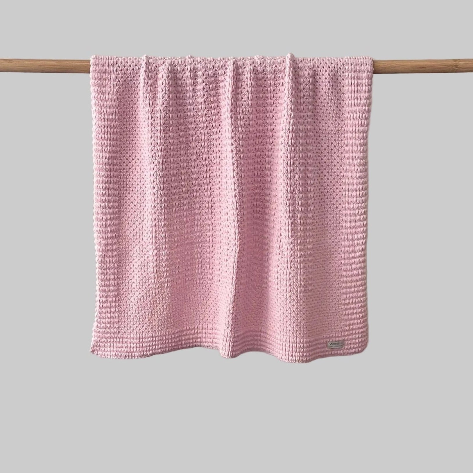 Textured blossom pink bassinet blanket hanging on a wooden rod against a grey background