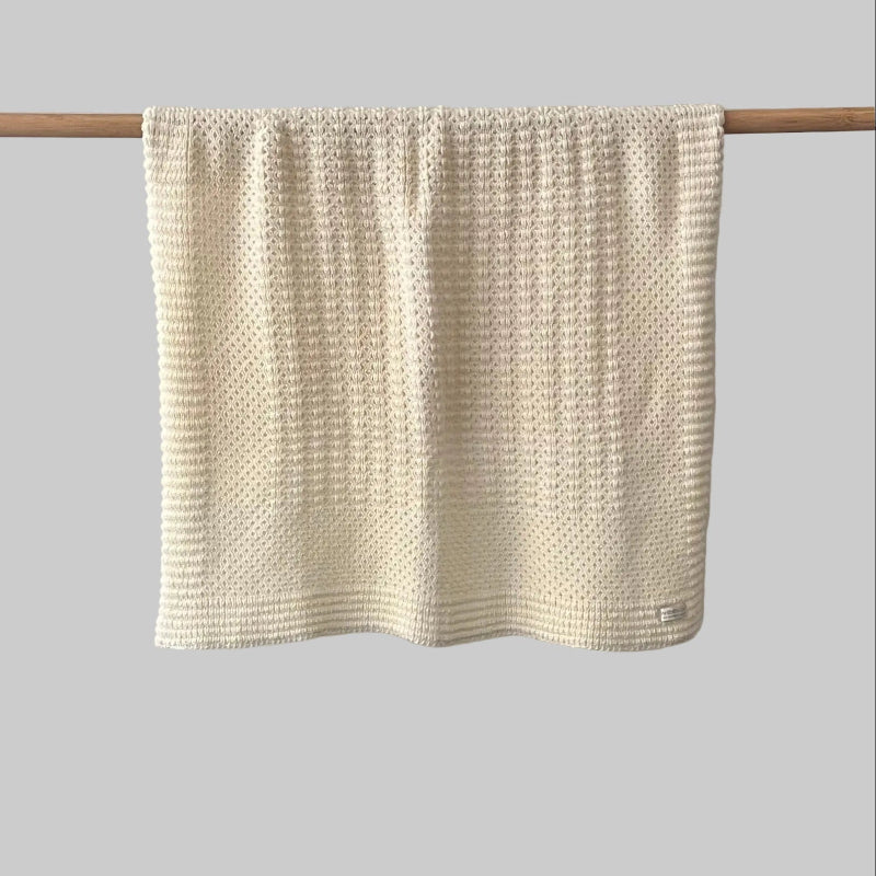 Textured buttermilk bassinet blanket hanging on a wooden rod against a grey background