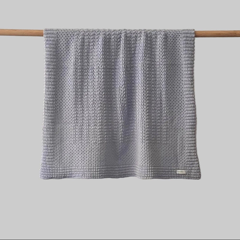 Textured misty blue bassinet blanket hanging on a wooden rod against a grey background