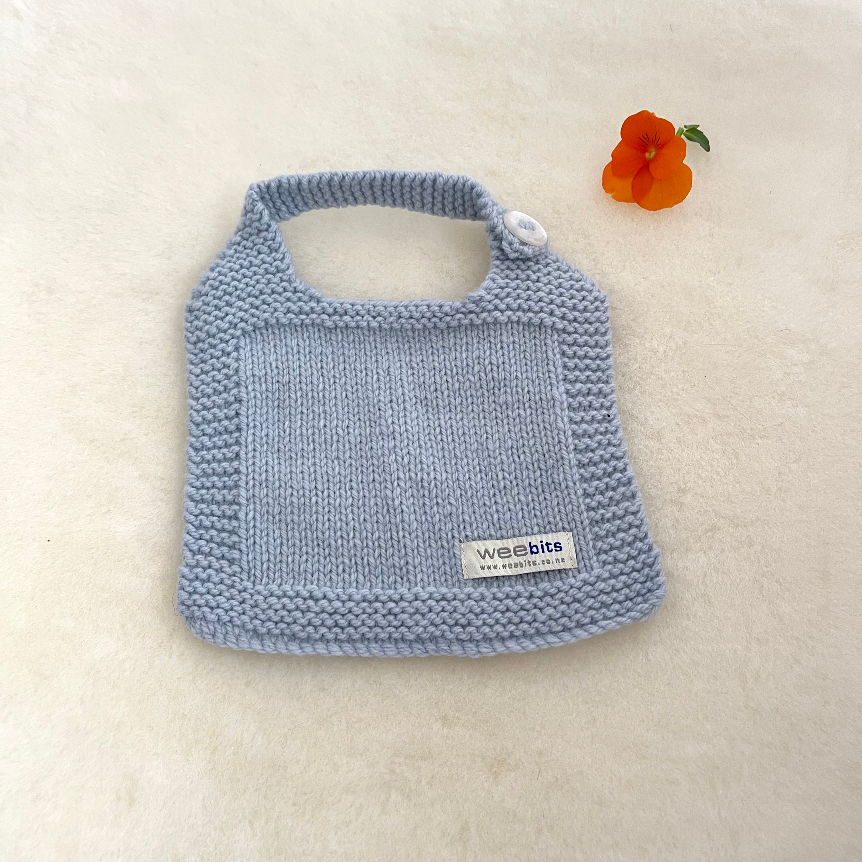 A light blue hand-knit baby bib with a button and a small 'weebits' brand label, laid flat on a cream-colored textured surface next to an orange flower.