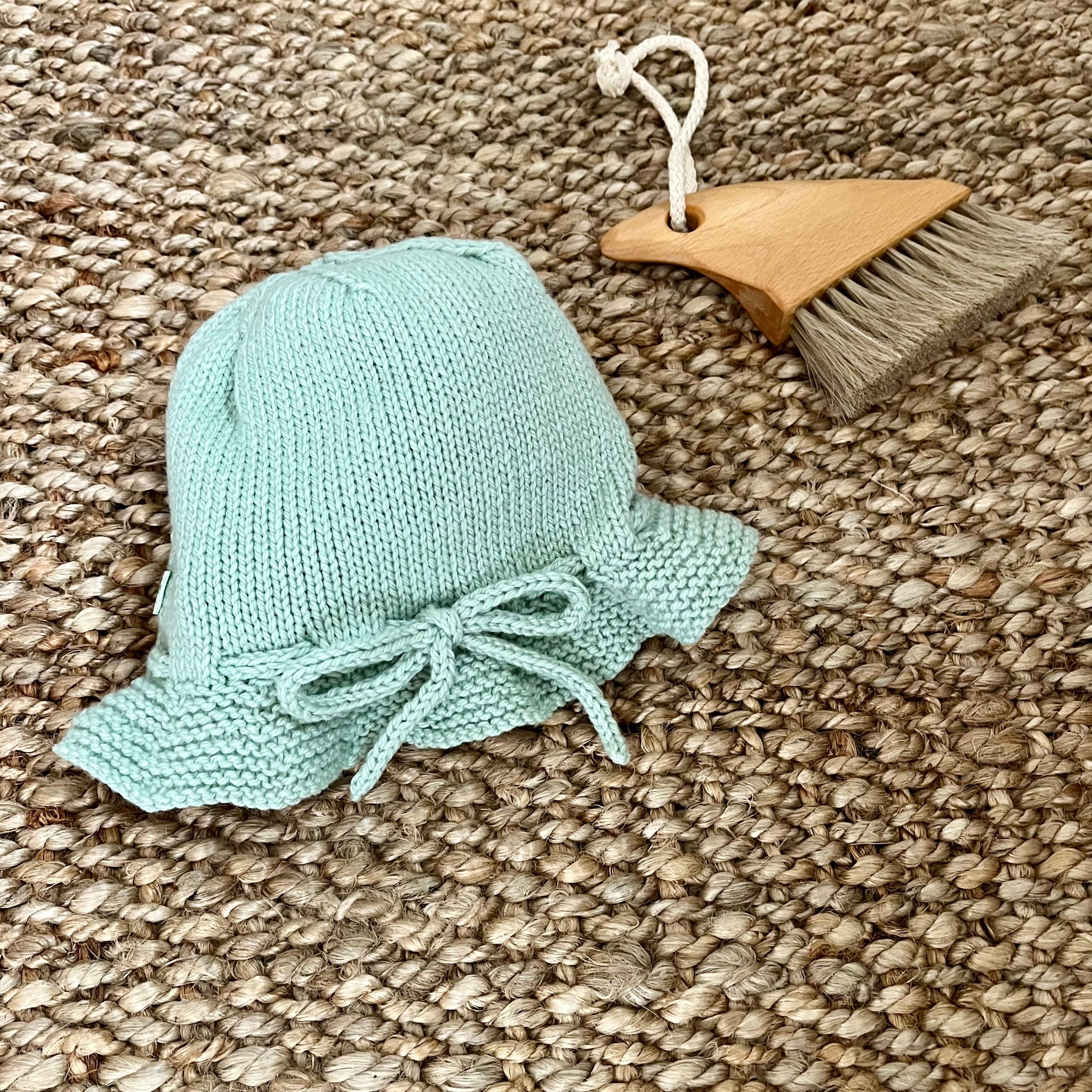 A small mint knitted bucket hat with a bow detail, displayed on a woven jute rug next to a wooden beach brush.