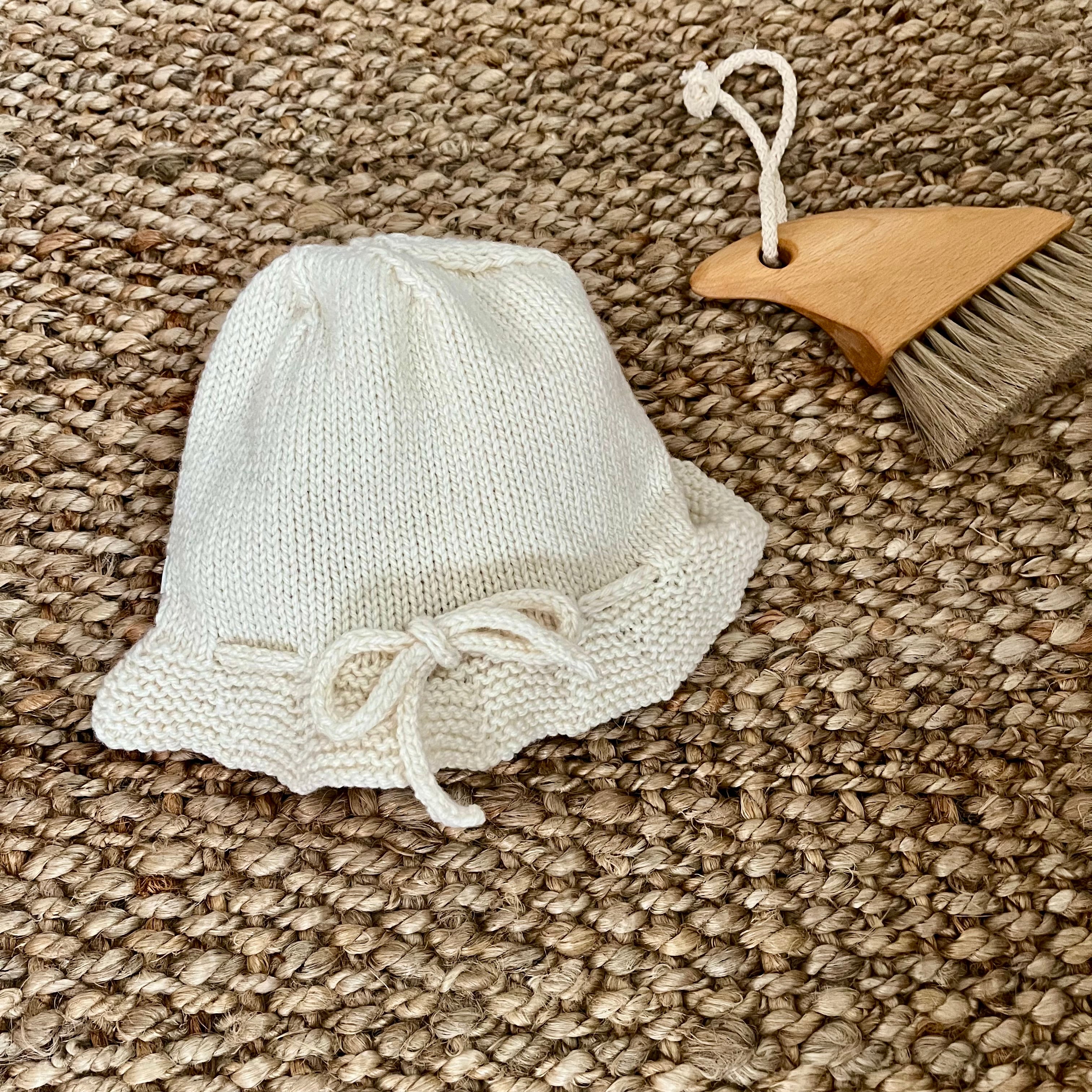 A small natural knitted bucket hat with a bow detail, displayed on a woven jute rug next to a wooden beach brush.