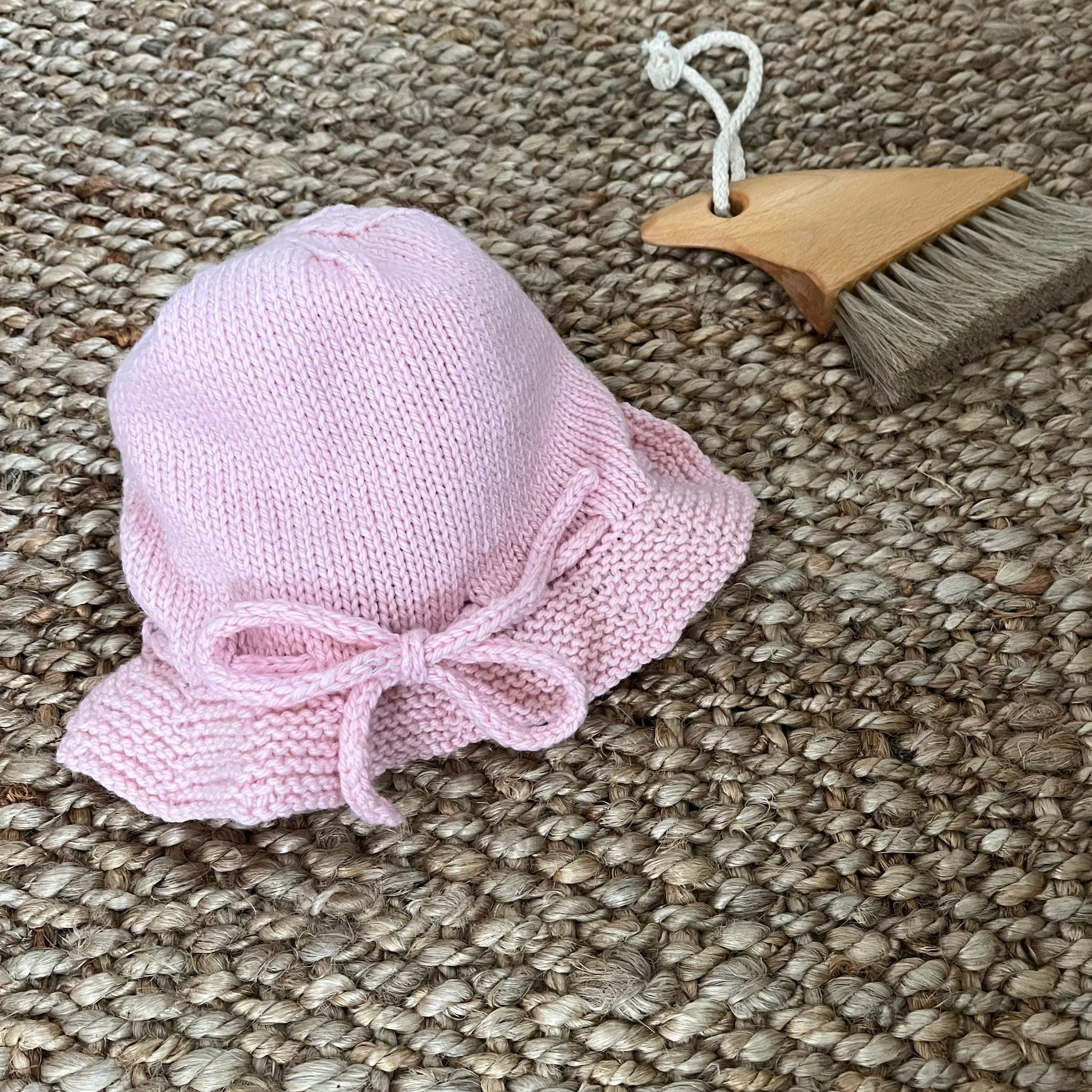 A small pink knitted bucket hat with a bow detail, displayed on a woven jute rug next to a wooden beach brush.