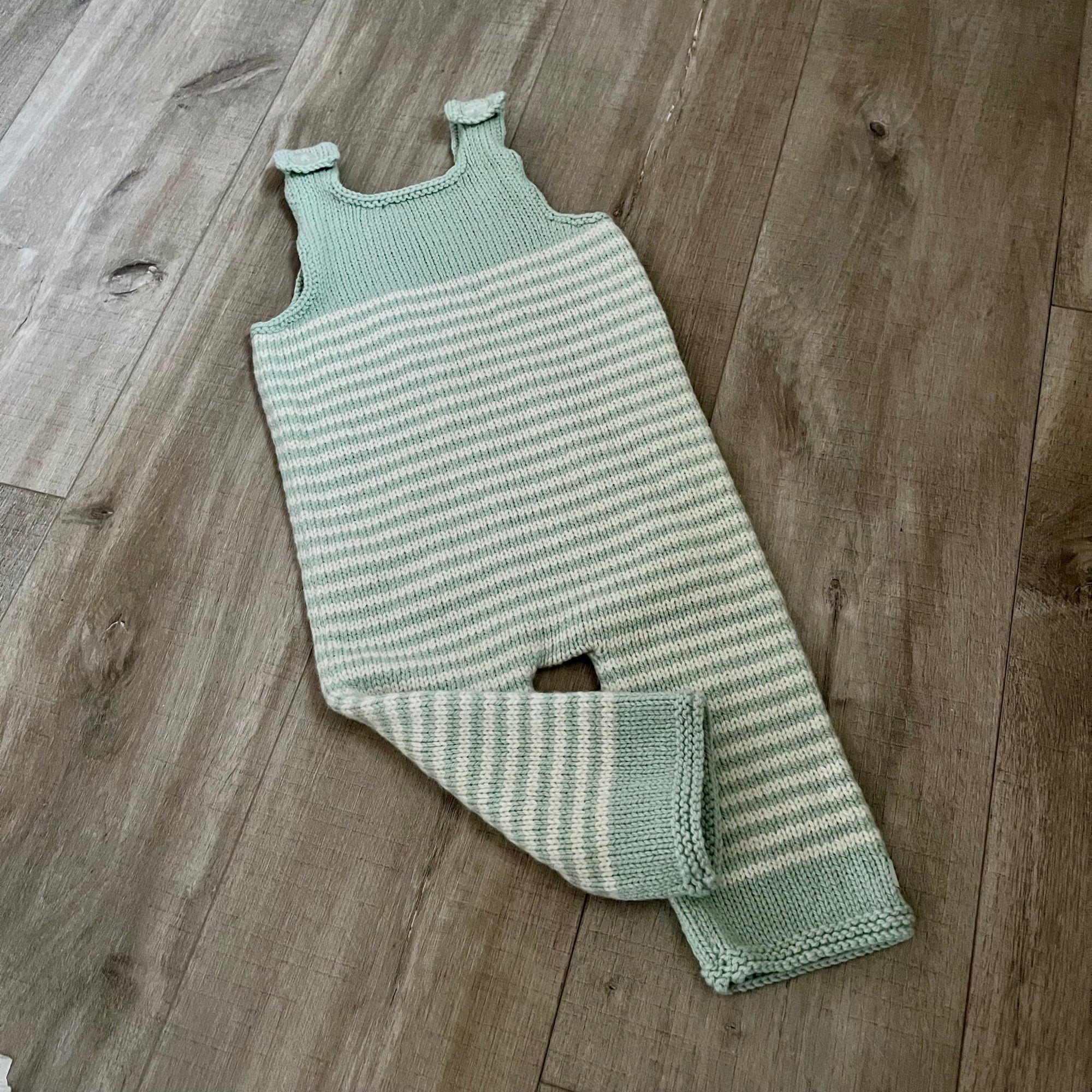 A mint natural stripe colored knit baby dungaree with shoulder straps and a leg opening, laid flat on a light brown wood floor.