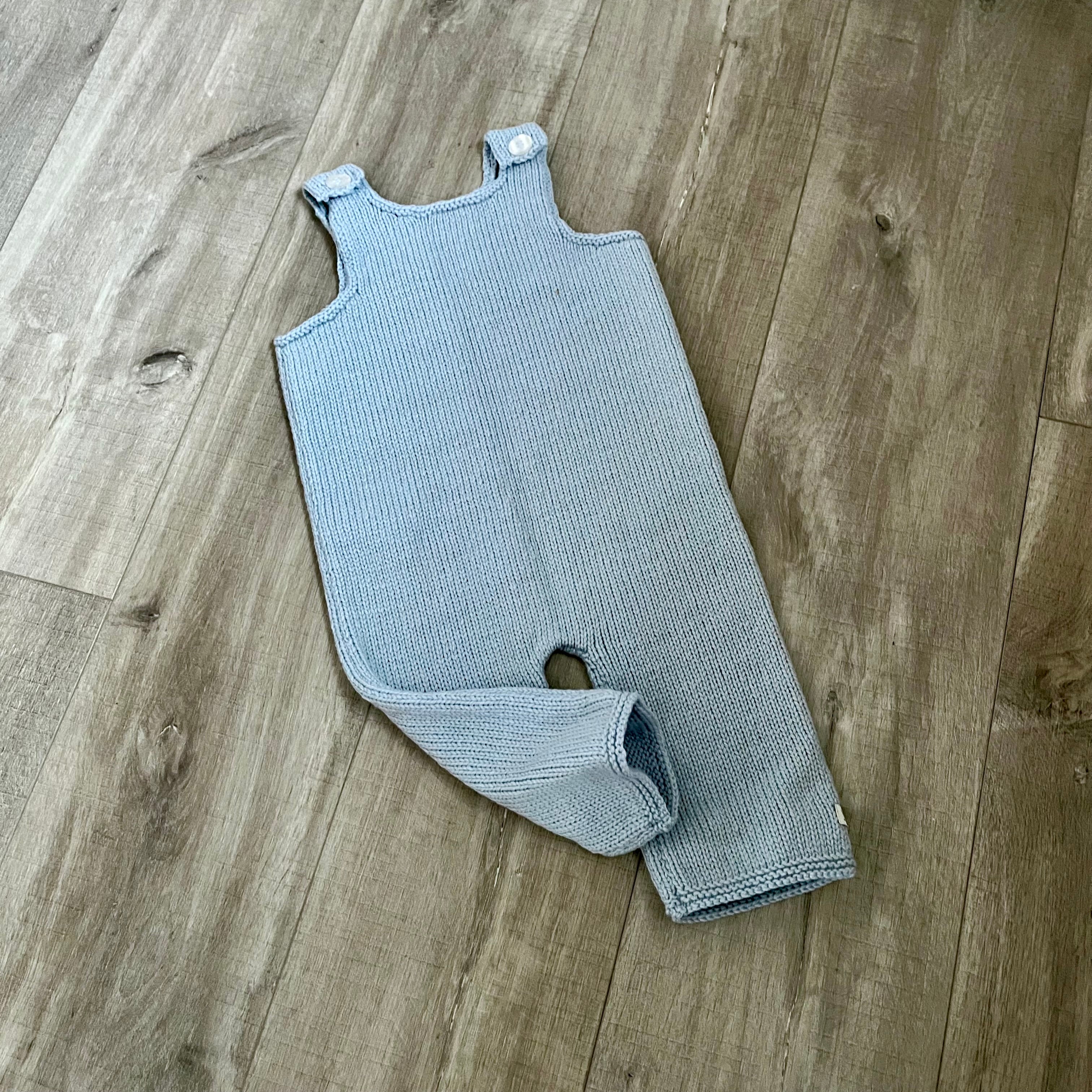A blue colored knit baby dungaree with shoulder straps and a leg opening, laid flat on a light brown wood floor.