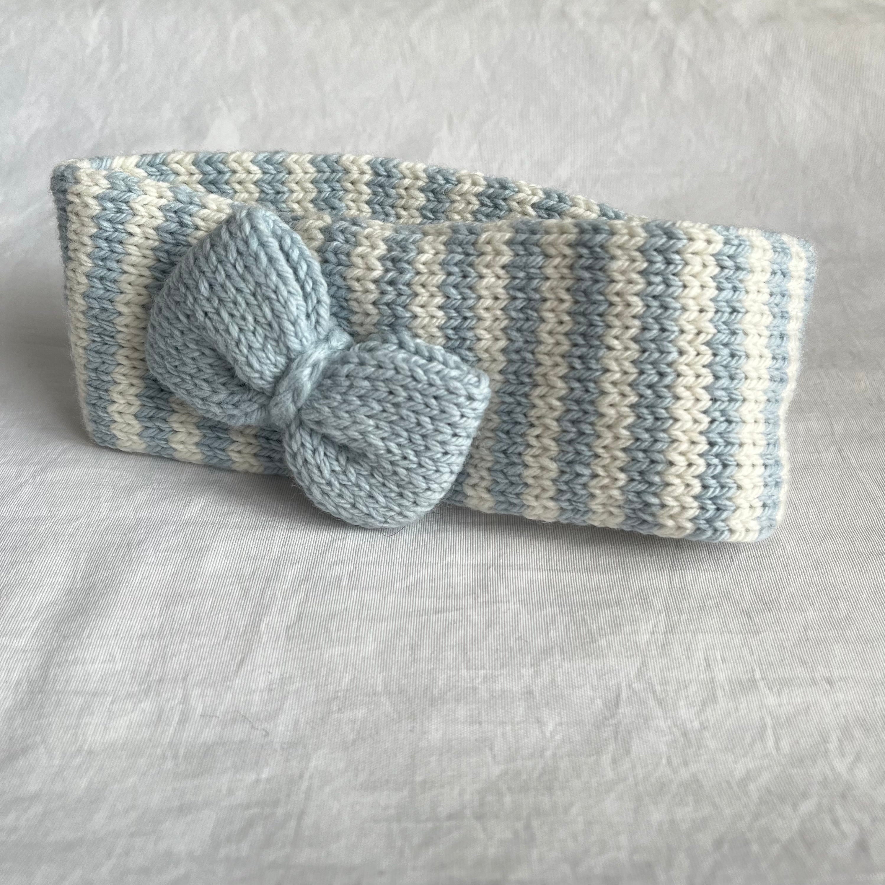 A handmade knit headband in a light blue and cream striped pattern, featuring a large matching knit bow on the front, displayed on a textured white fabric background.