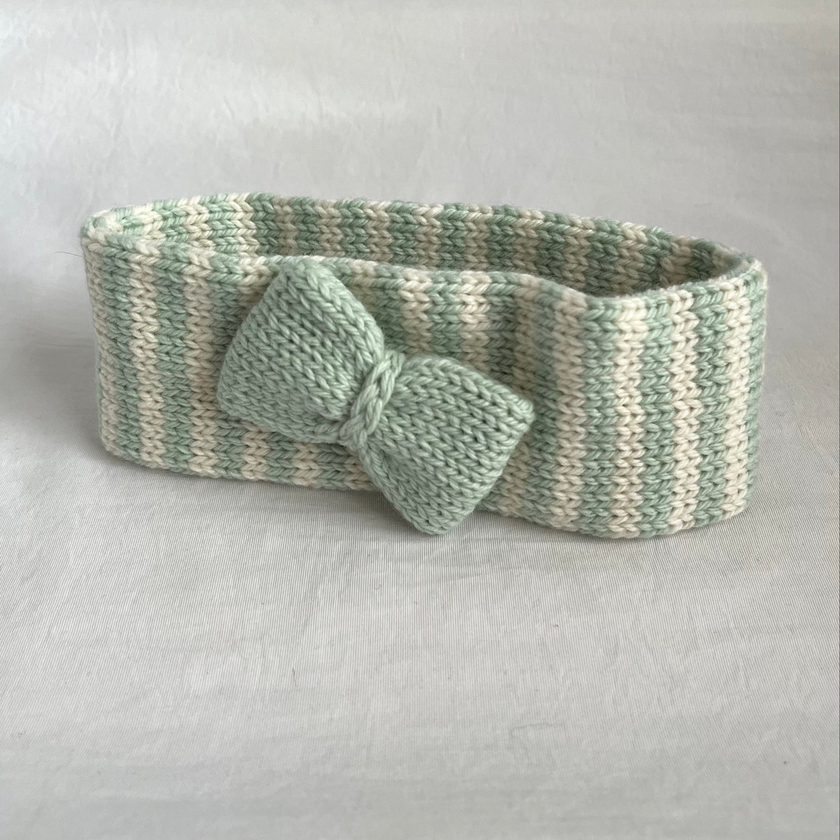 A handmade knit headband with a matching bow, featuring a striped pattern of sage green and cream yarn, set against a neutral light gray background.