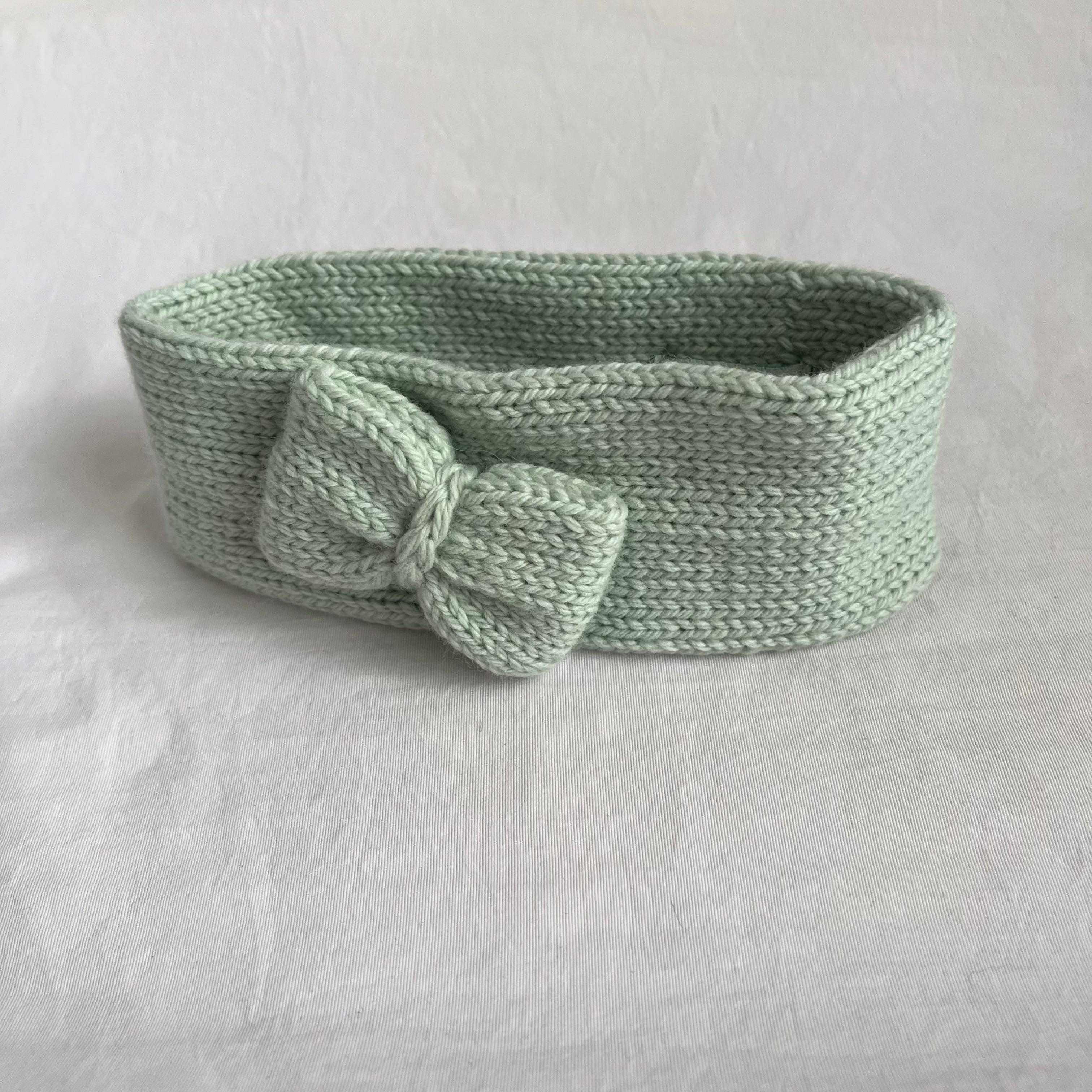A light sage green knitted headband with a large bow detail on the front, displayed on a textured white fabric background.