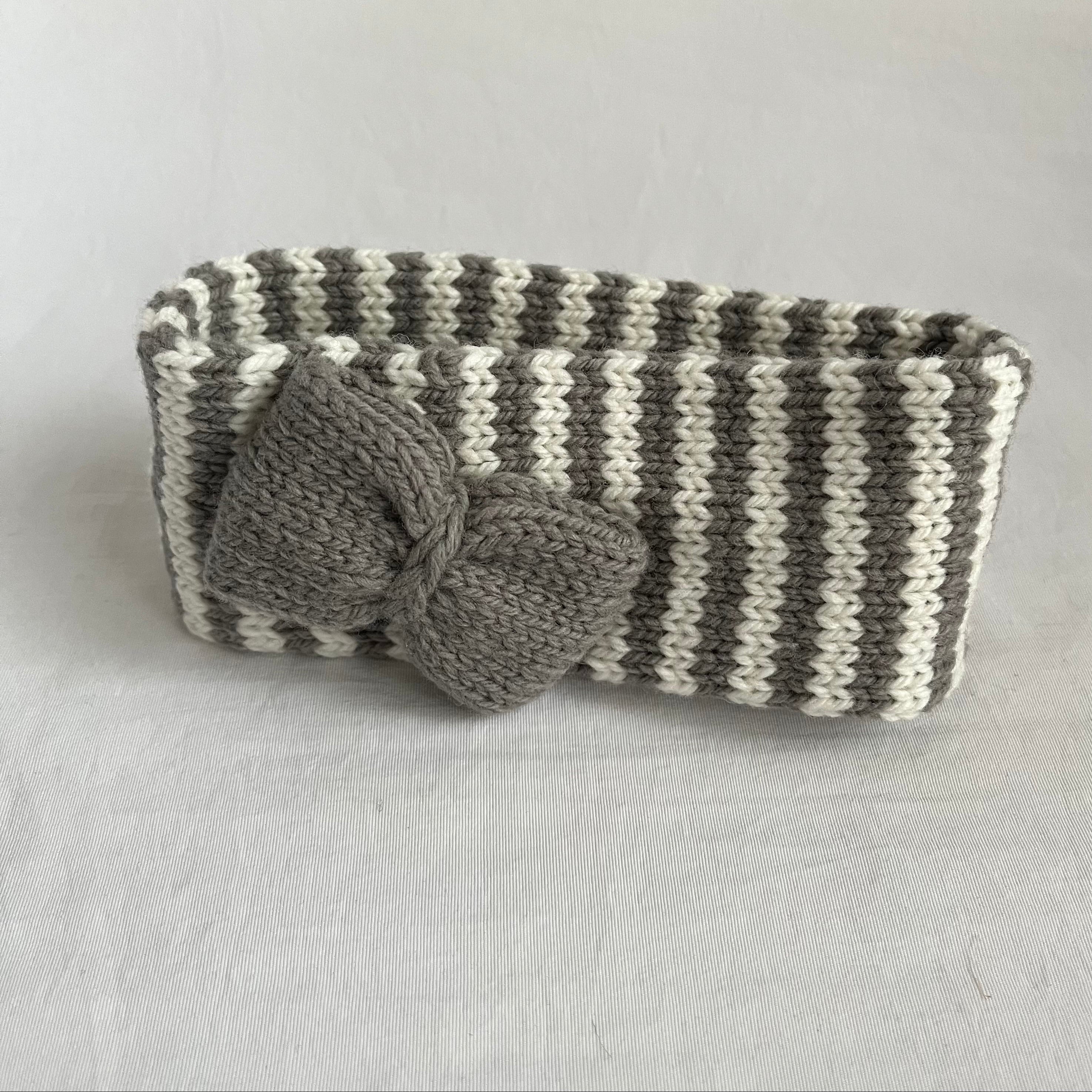 A handmade knit headband with a large gray bow, featuring alternating gray and off-white stripes, set against a neutral light gray background.