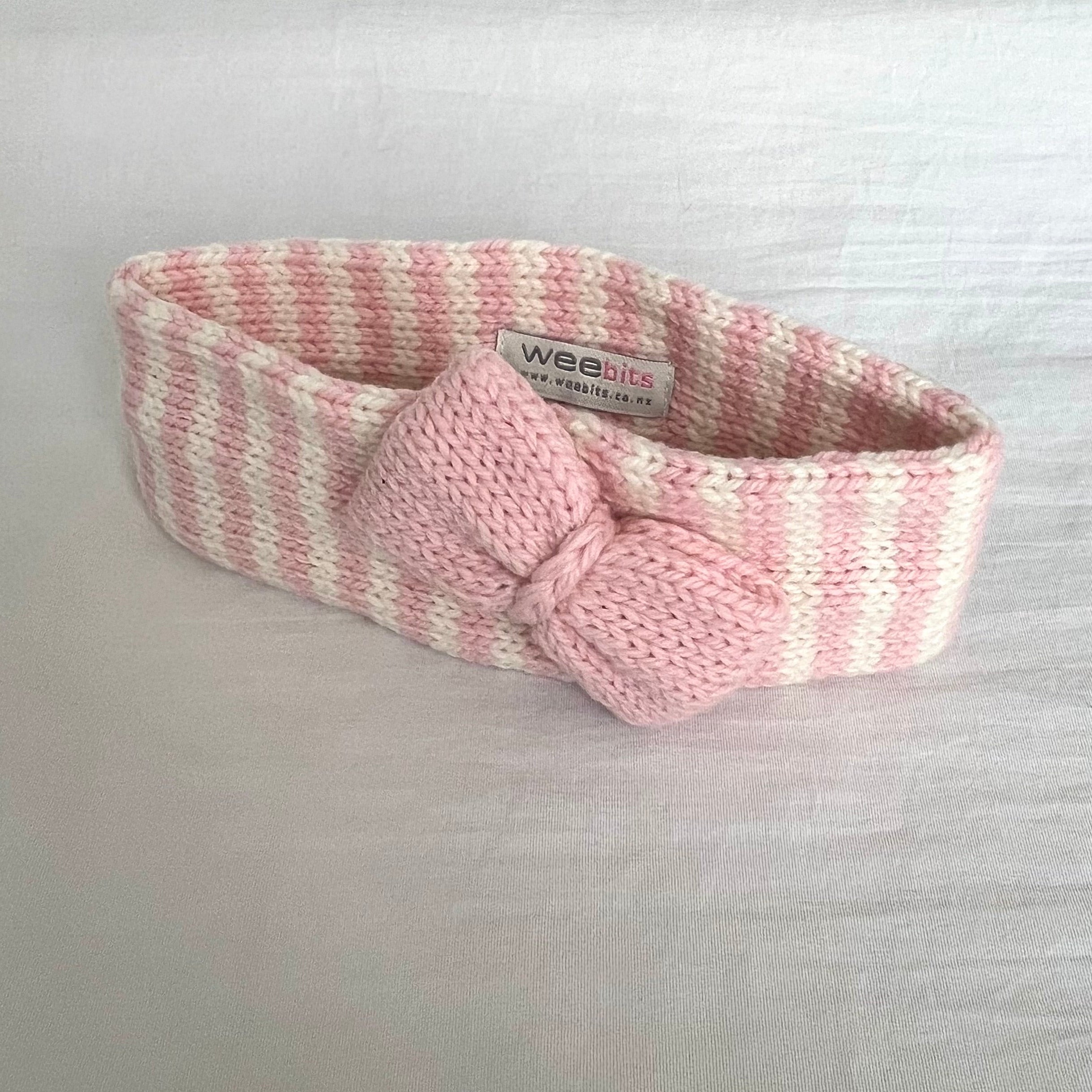 A pink and white striped knit headband with a matching knotted bow, shown on a light gray fabric background.