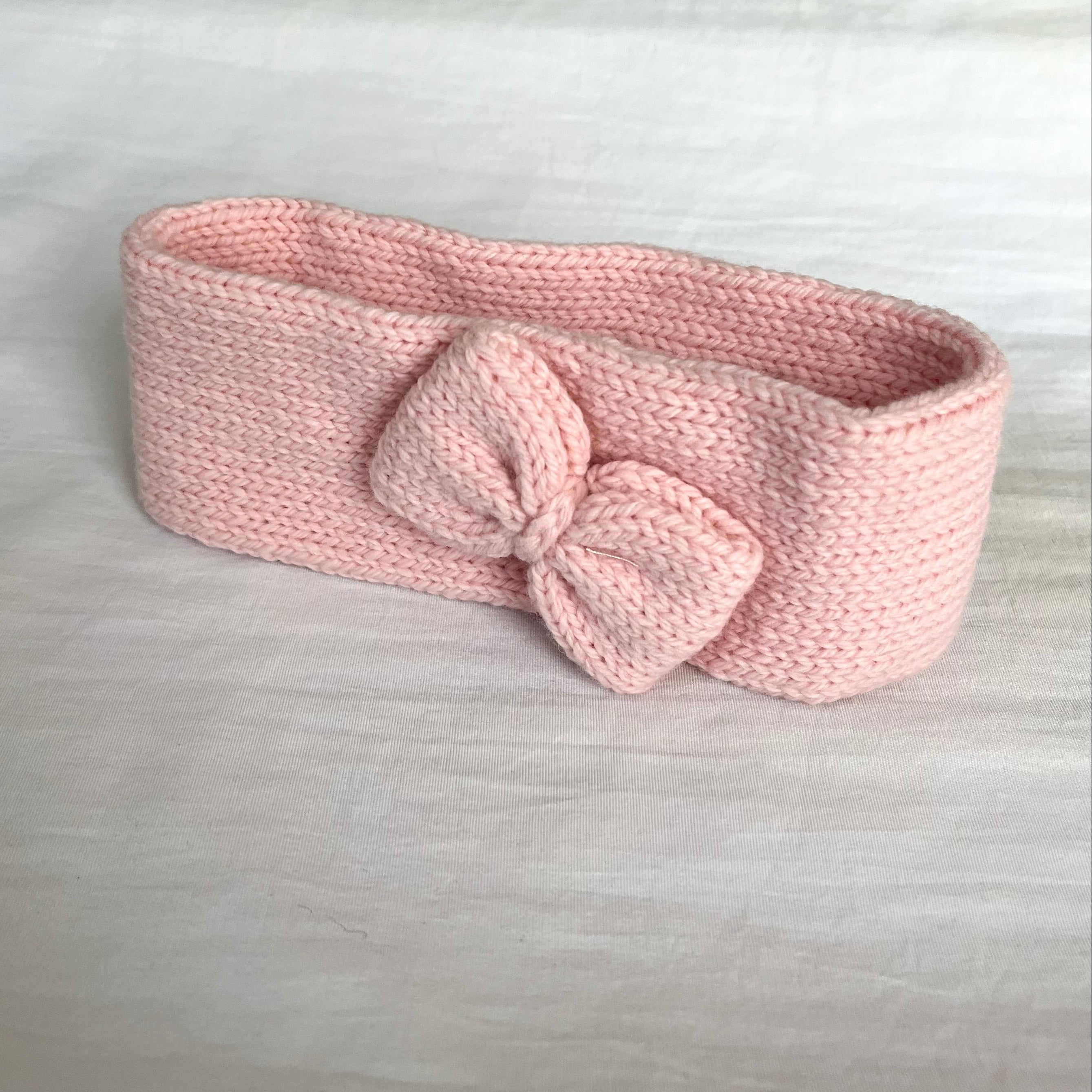 A light pink knitted headband with a matching bow, displayed on a white fabric surface.