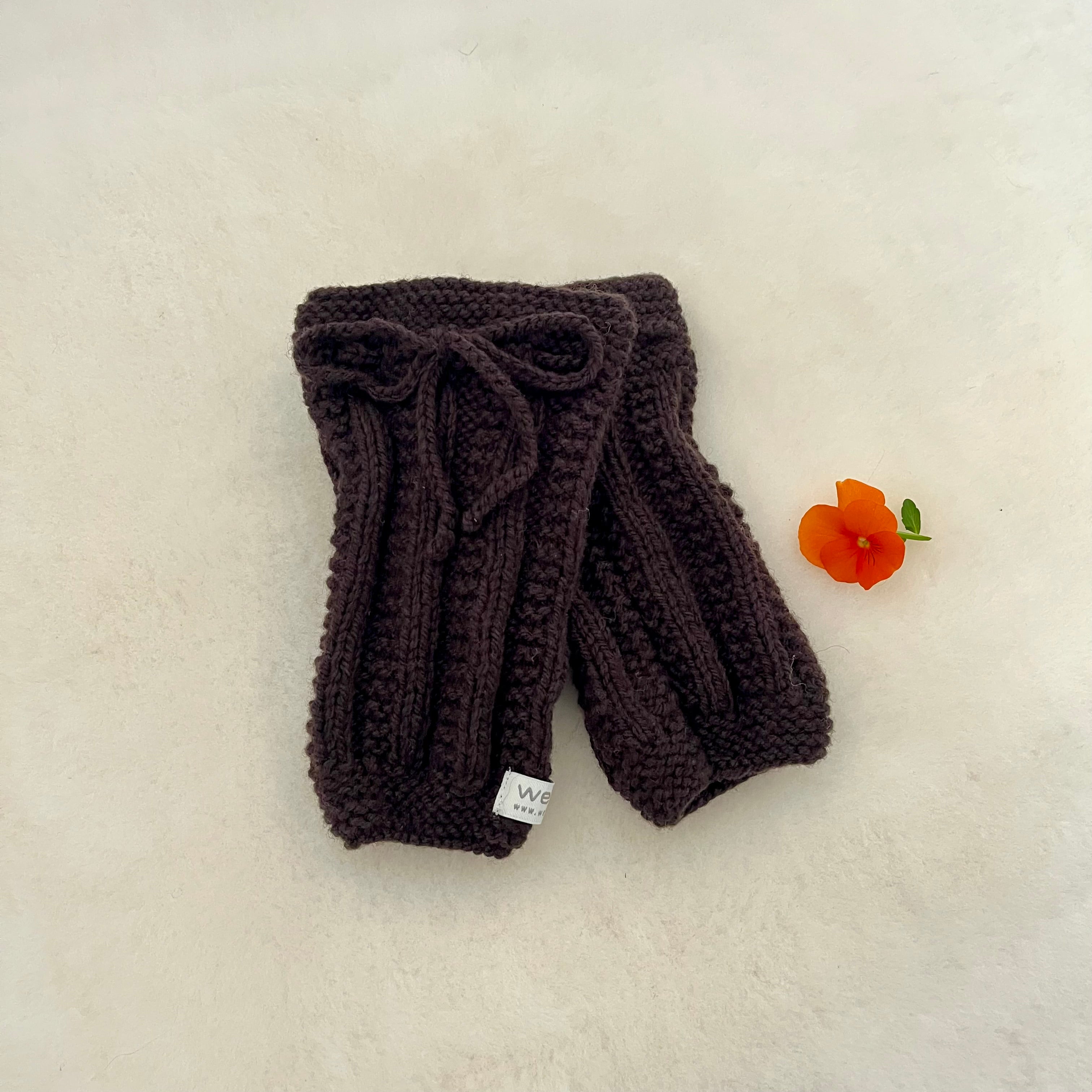A pair of chocolate hand-knitted legwarmers with a matching knitted bow on the top, displayed on a textured off-white surface next to a small orange flower.