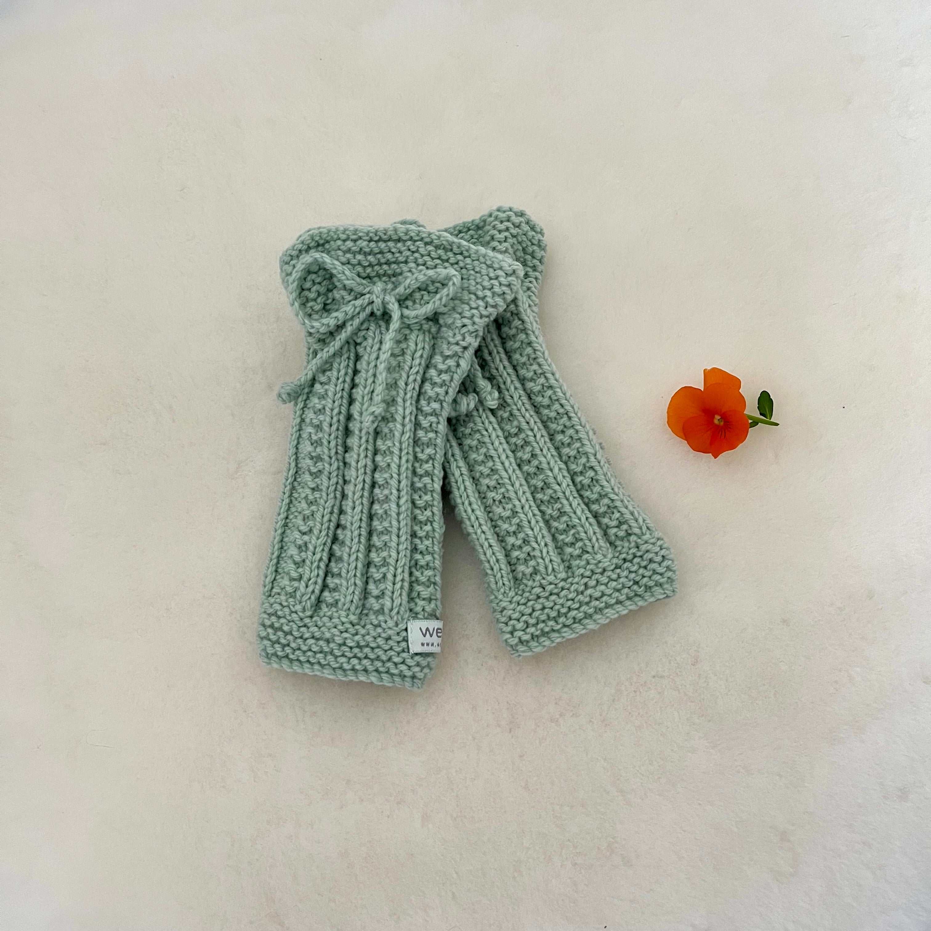 A pair of pale sage green hand-knitted legwarmers with a matching knitted bow on the top, displayed on a textured off-white surface next to a small orange flower.