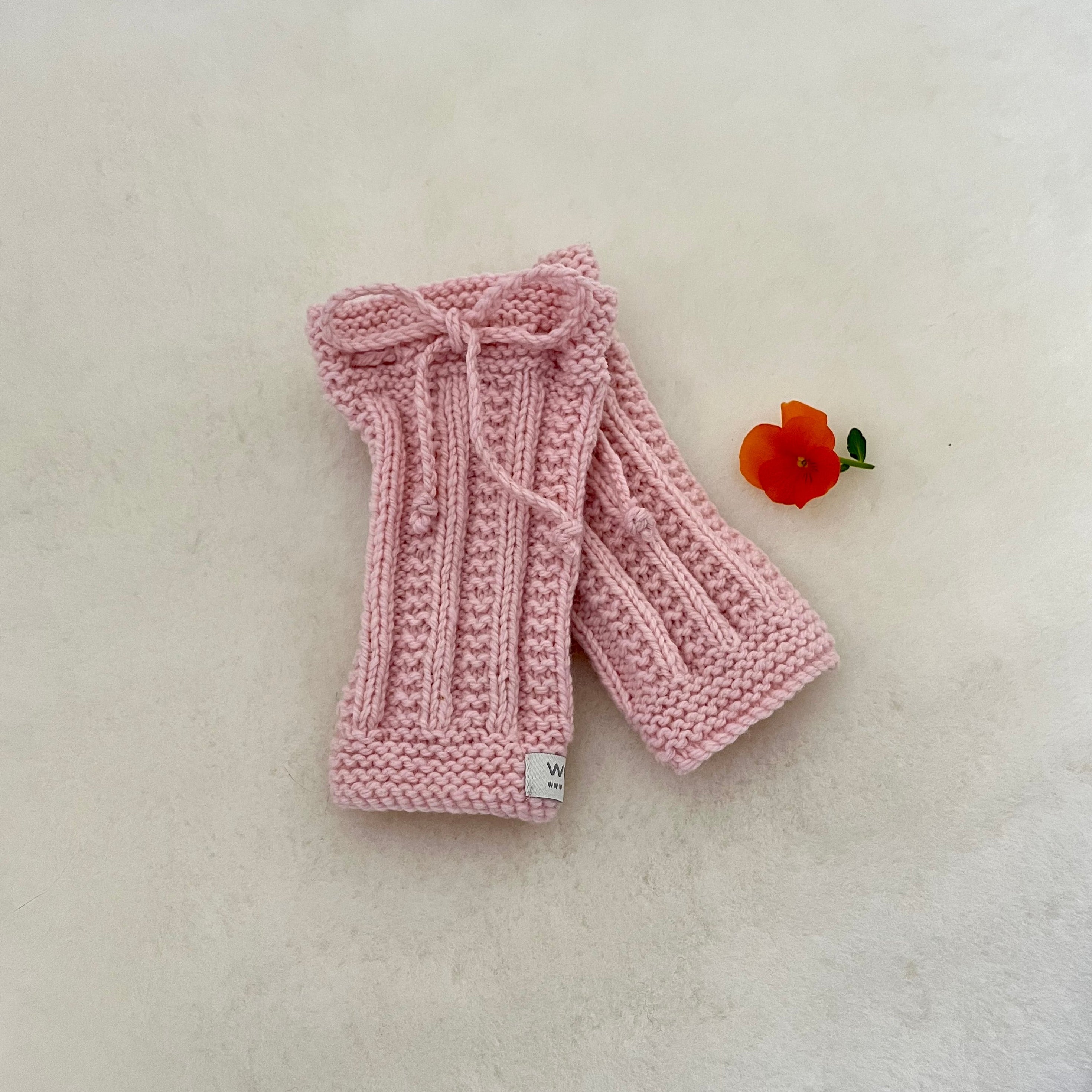 A pair of pink hand-knitted legwarmers with a matching knitted bow on the top, displayed on a textured off-white surface next to a small orange flower.