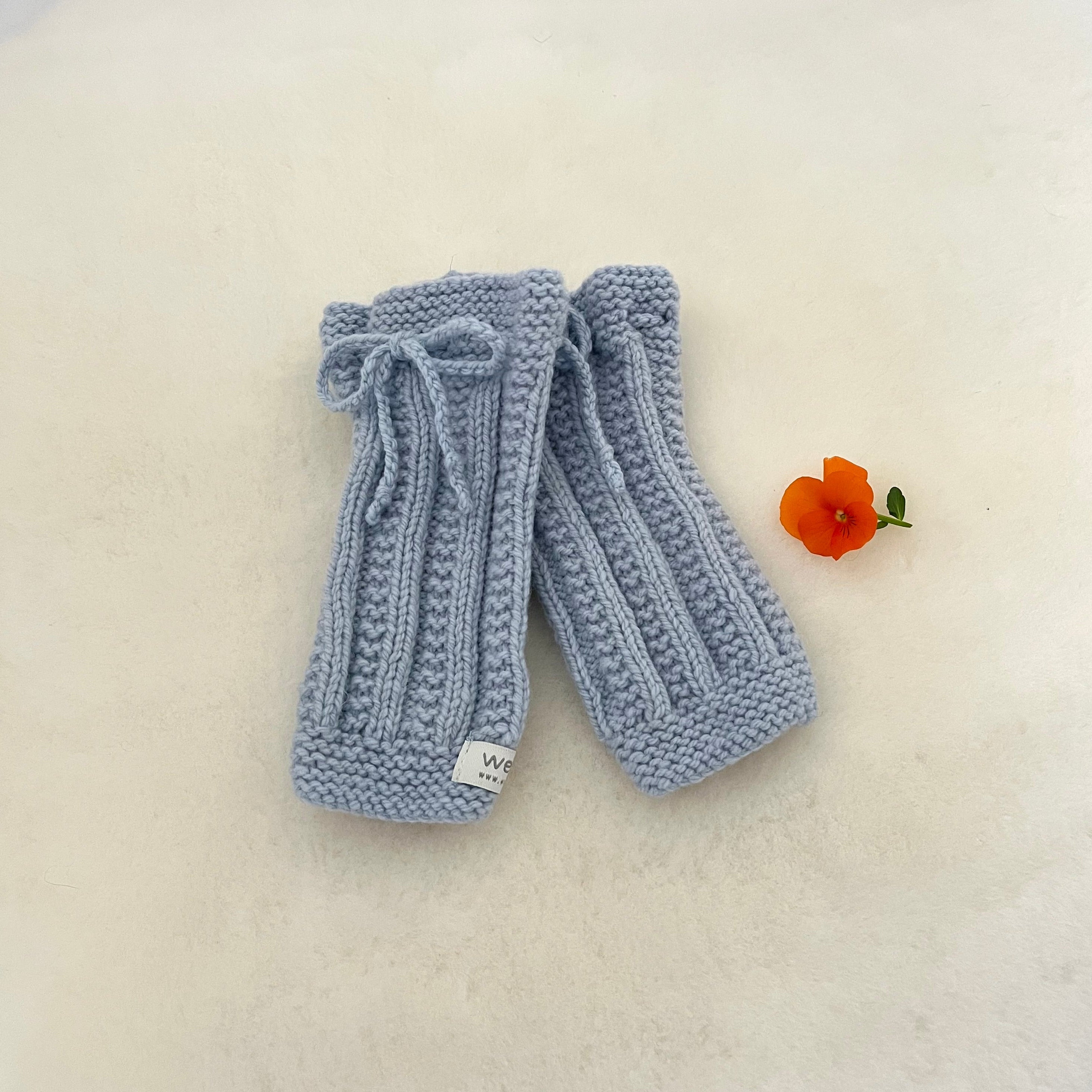 A pair of powder blue hand-knitted legwarmers with a matching knitted bow on the top, displayed on a textured off-white surface next to a small orange flower.