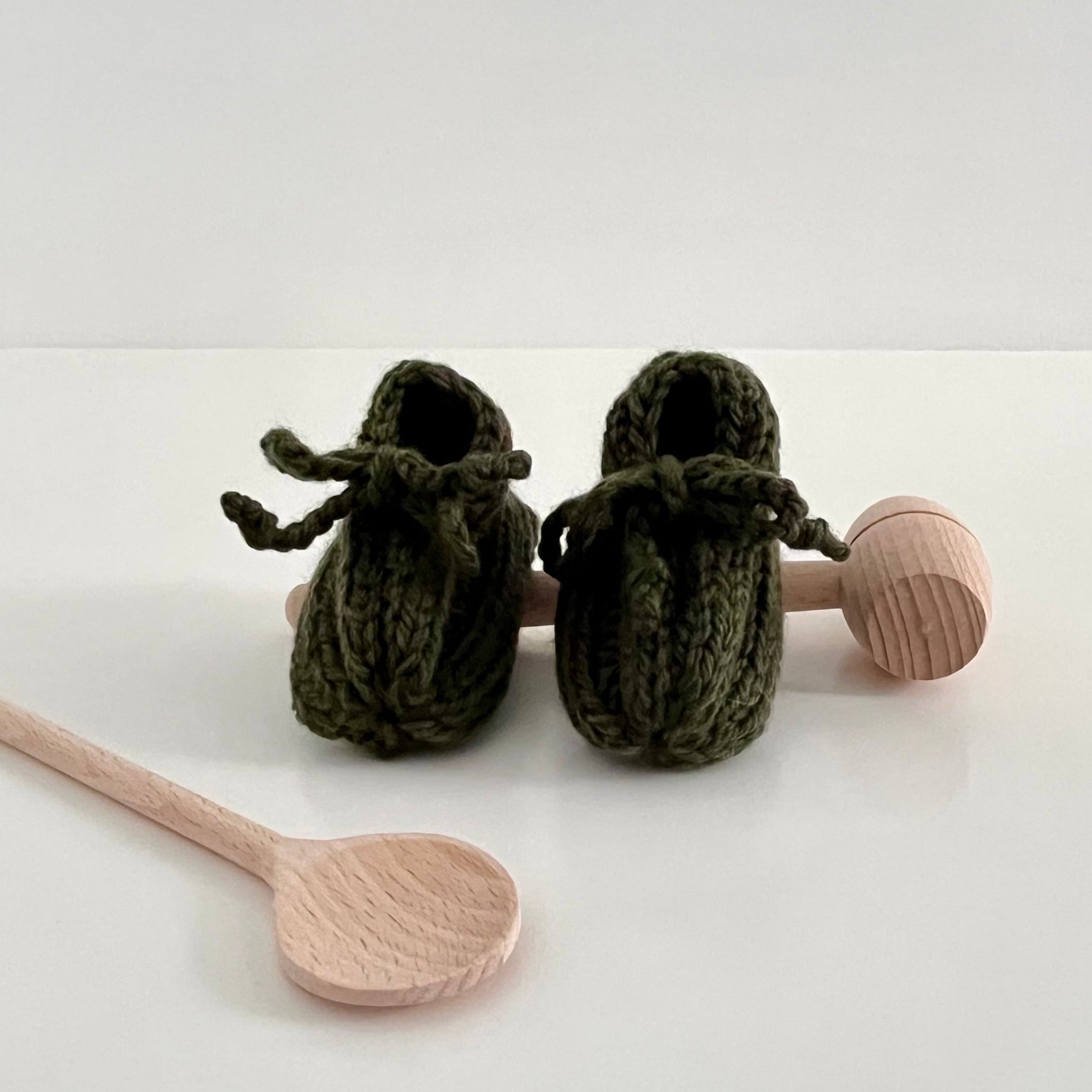 Forest green knitted baby booties with wooden rattle and spoon on a white background