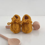 Pair of mustard knitted baby booties on a white background with wooden spoons for scale.