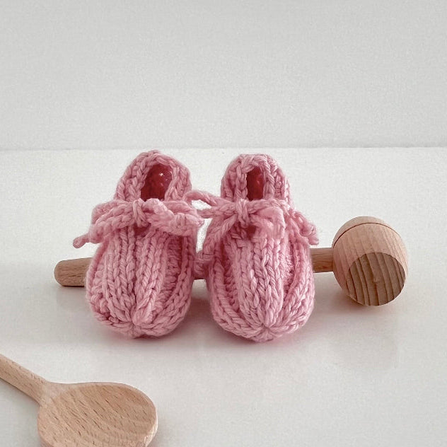 Pair of pink knitted baby booties with wooden spoons on a white background