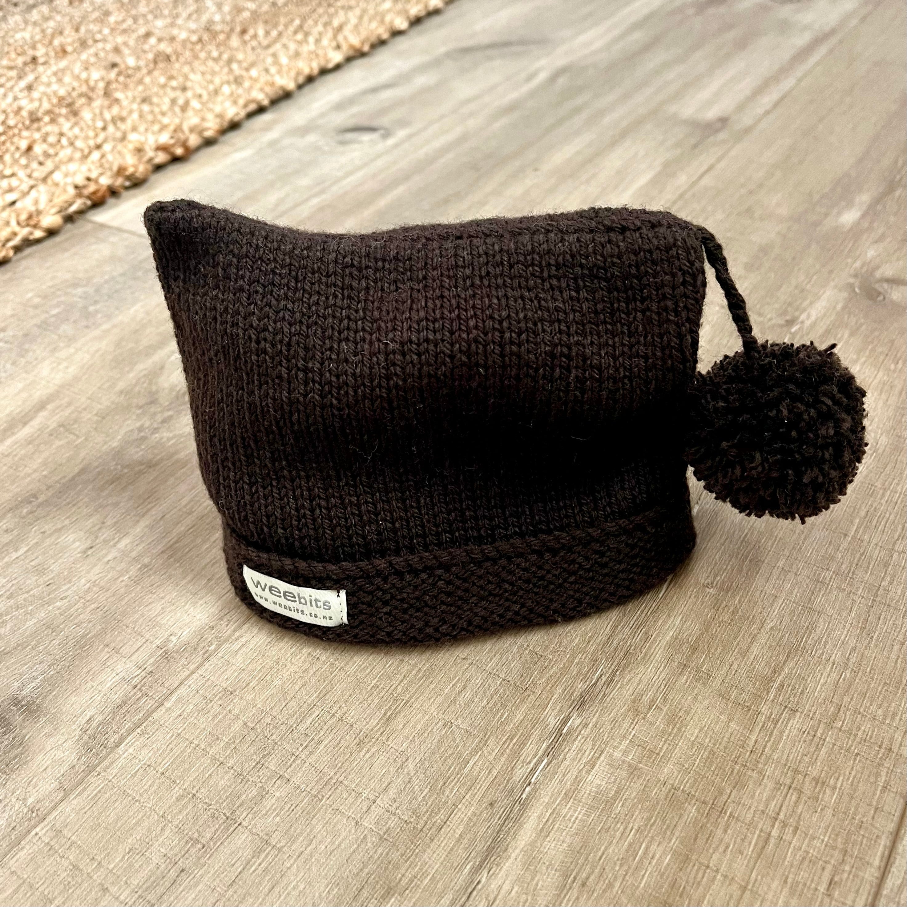 A dark brown knit beanie hat with a matching pom-pom and a small white brand label reading 'weebits', placed on a light wood floor.