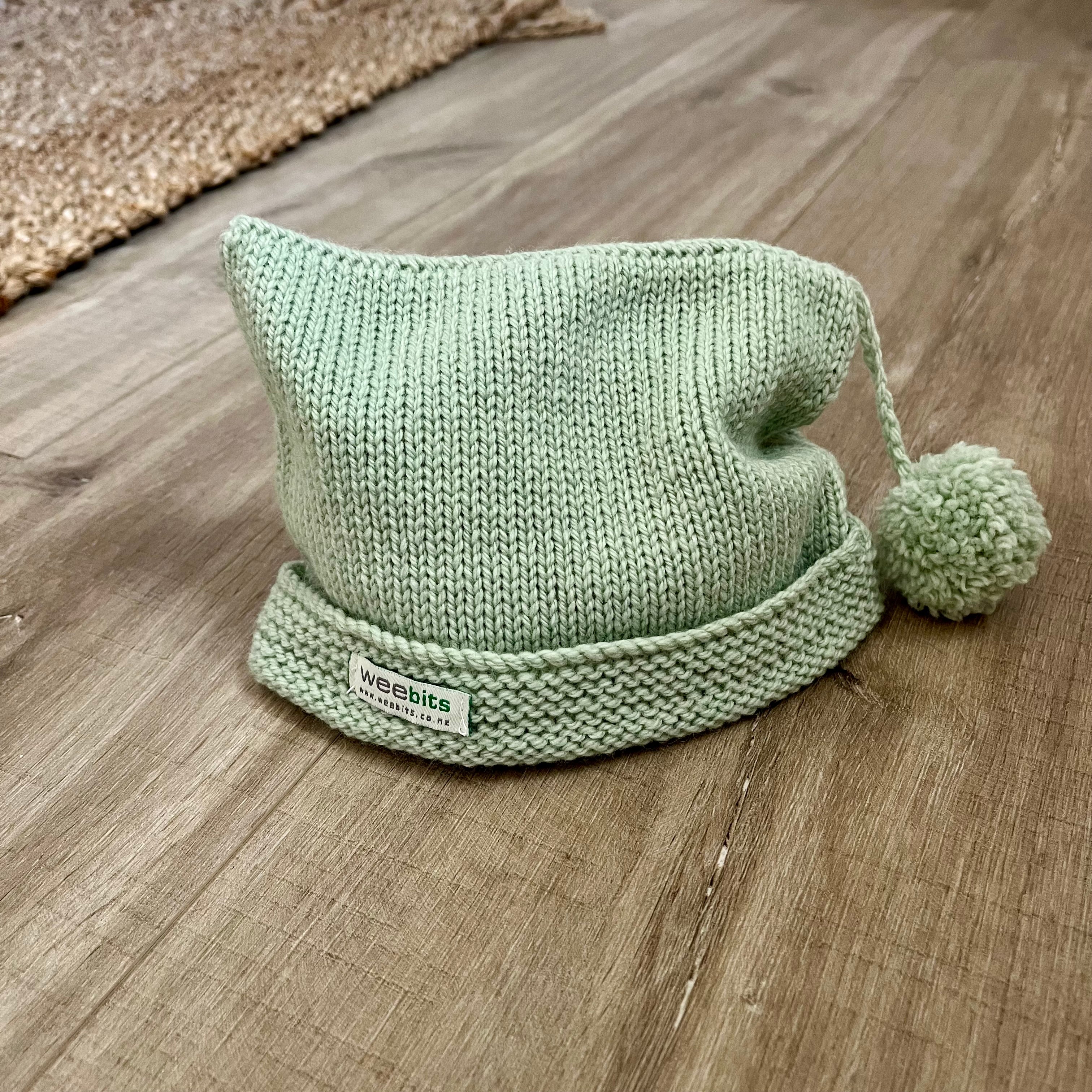 A light sage green knit beanie hat with a folded cuff and a matching pom-pom, resting on a wooden floor. A small white brand label reading 'weebits' is visible on the cuff.