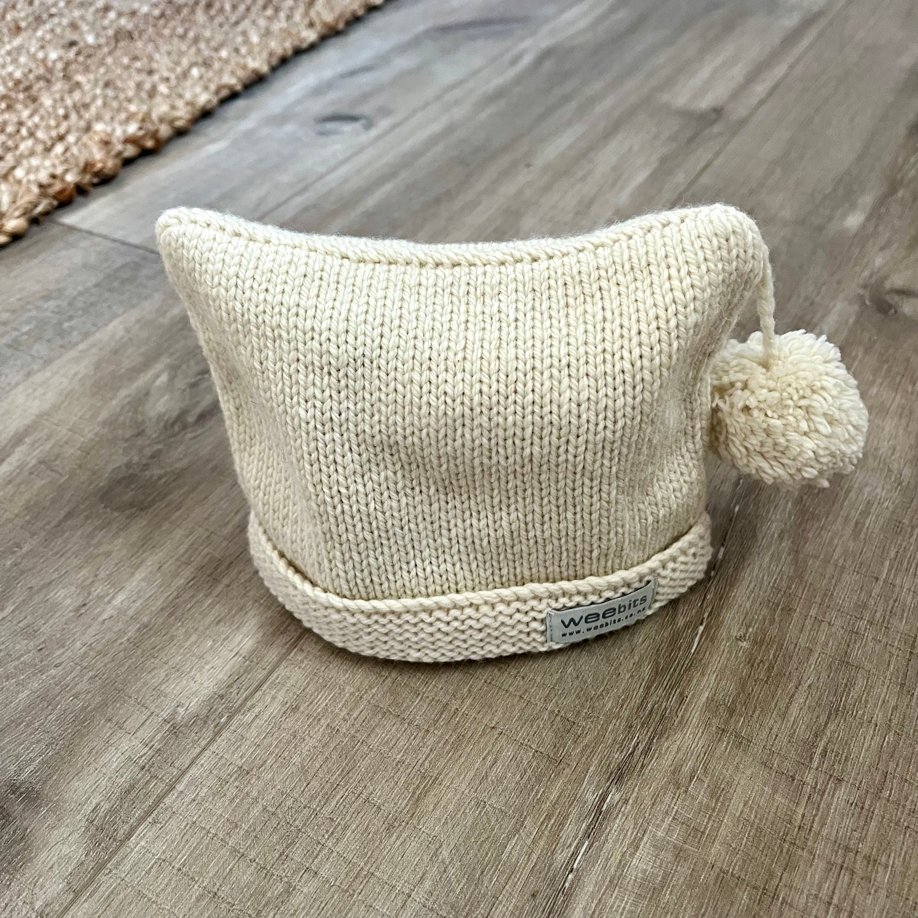 A cream-colored knit beanie hat with a matching pom-pom on the side on brown wood floor.