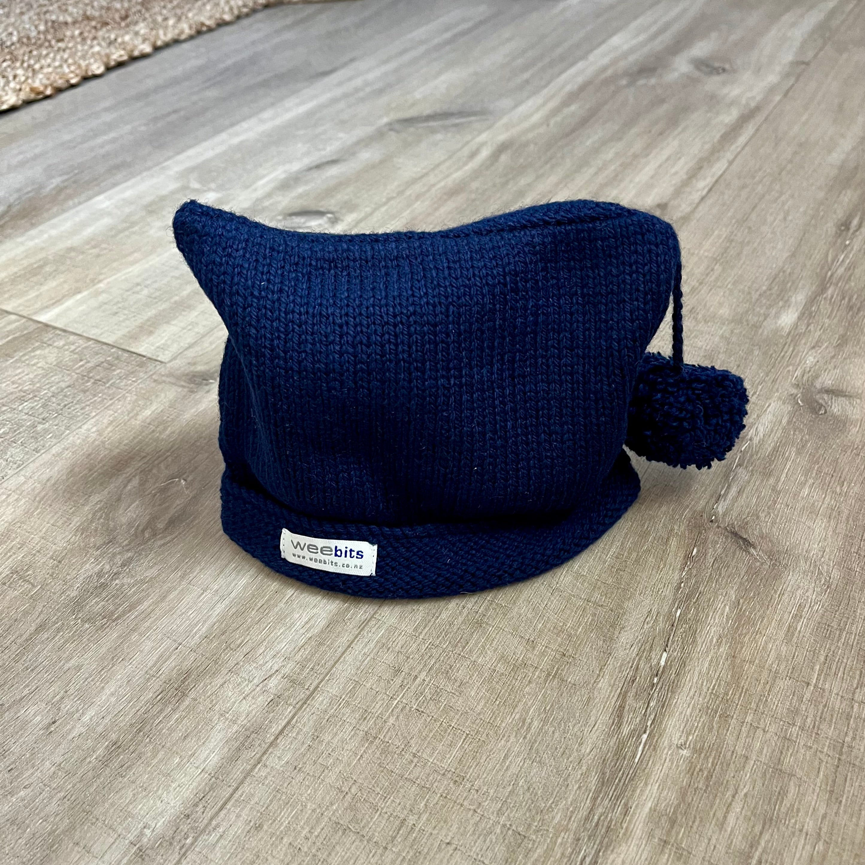 A navy blue knit cat-ear beanie hat with a pom-pom and a small white 'weebits' brand label, resting on a light wood floor.