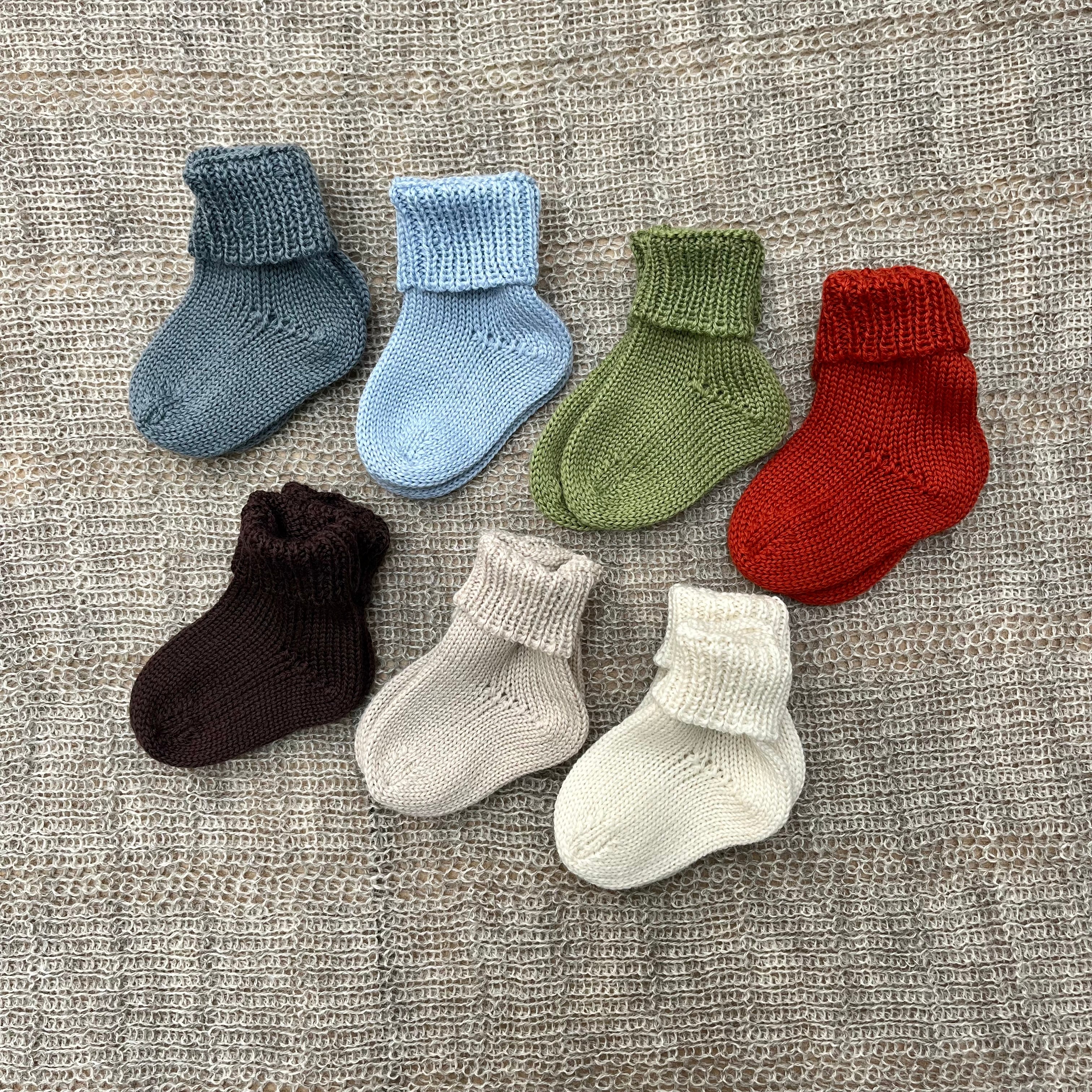 Six pairs of hand-knit baby socks in assorted colors, including blue, green, red, brown, cream, and light blue, arranged on a textured beige woven blanket.