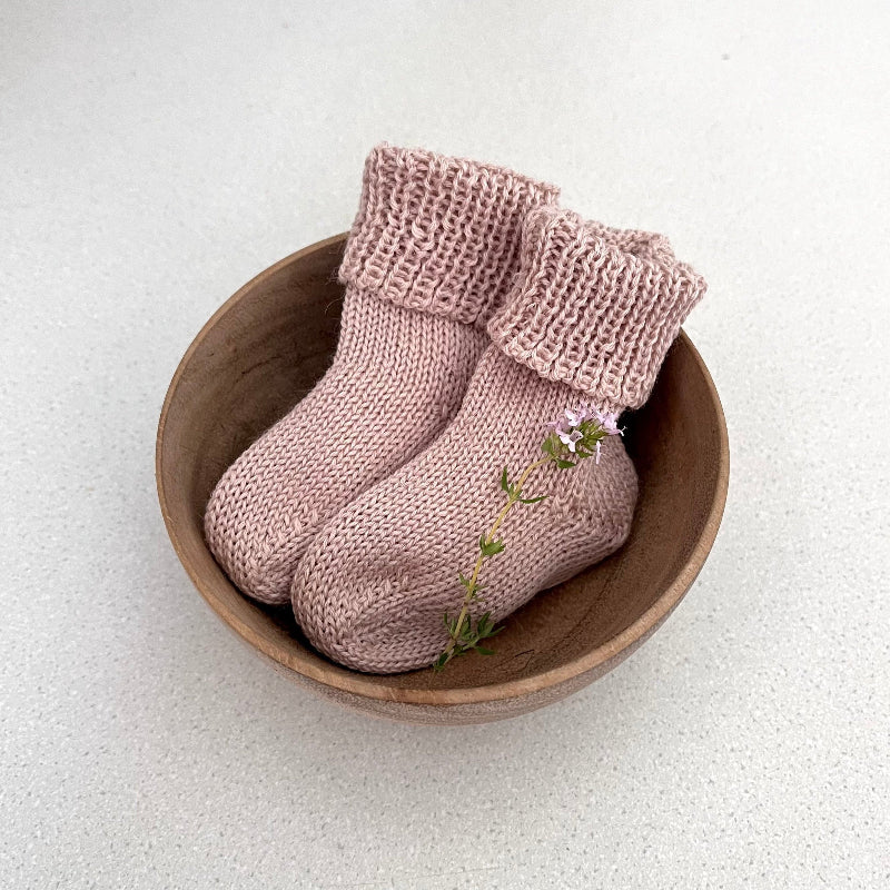 Pair of warm sandstone baby socks in a wooden bowl on a light background