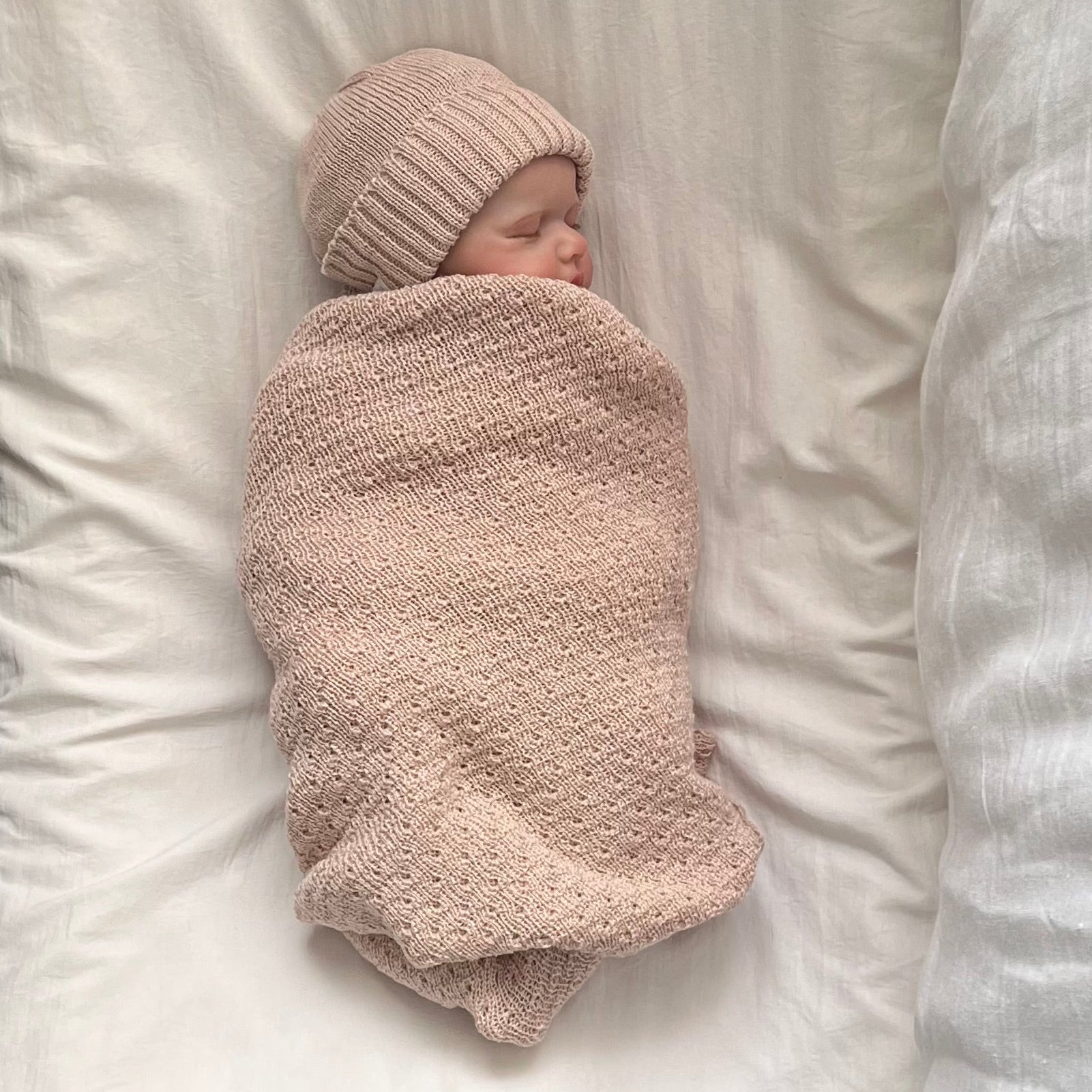 A newborn baby swaddled in a sandstone textured blanket and wearing a matching knit beanie, lying on a white pillow between white and gray bedding.