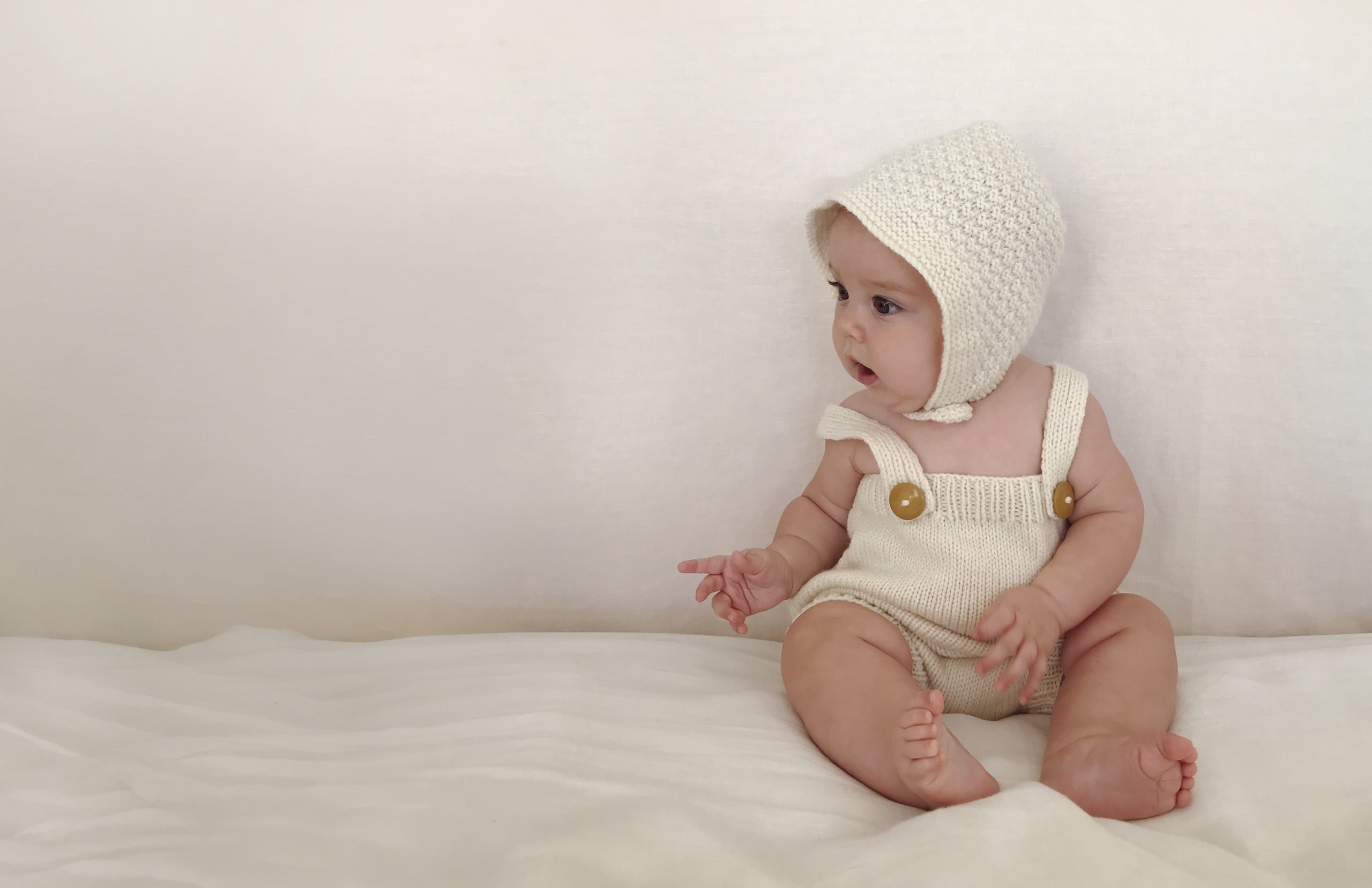 A baby wearing a cream knit bonnet and matching cream overalls with gold buttons, sitting on a white blanket against a white background.