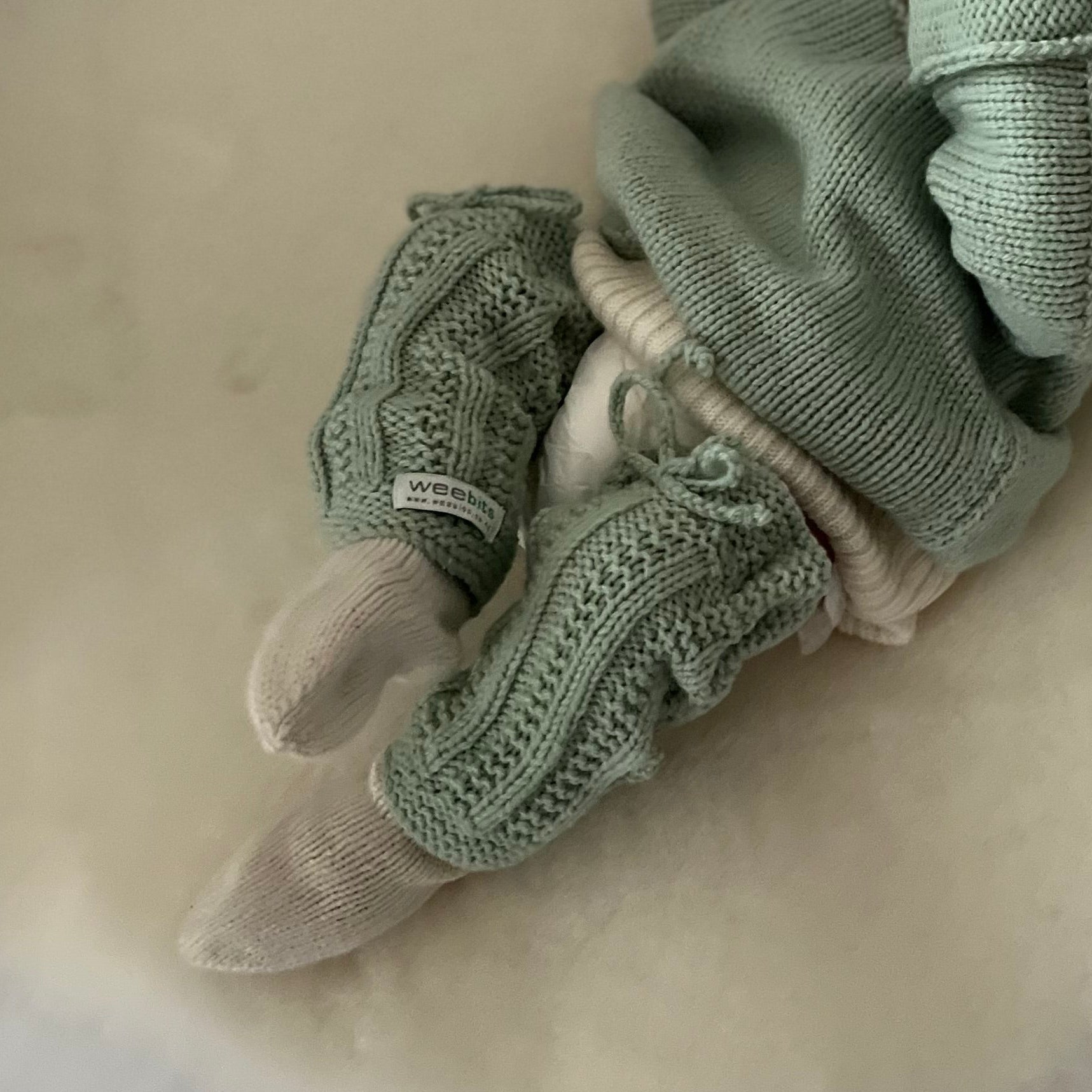 A baby dressed in a pale sage green knit outfit, lying on a white sheepskin rug on a white sofa.