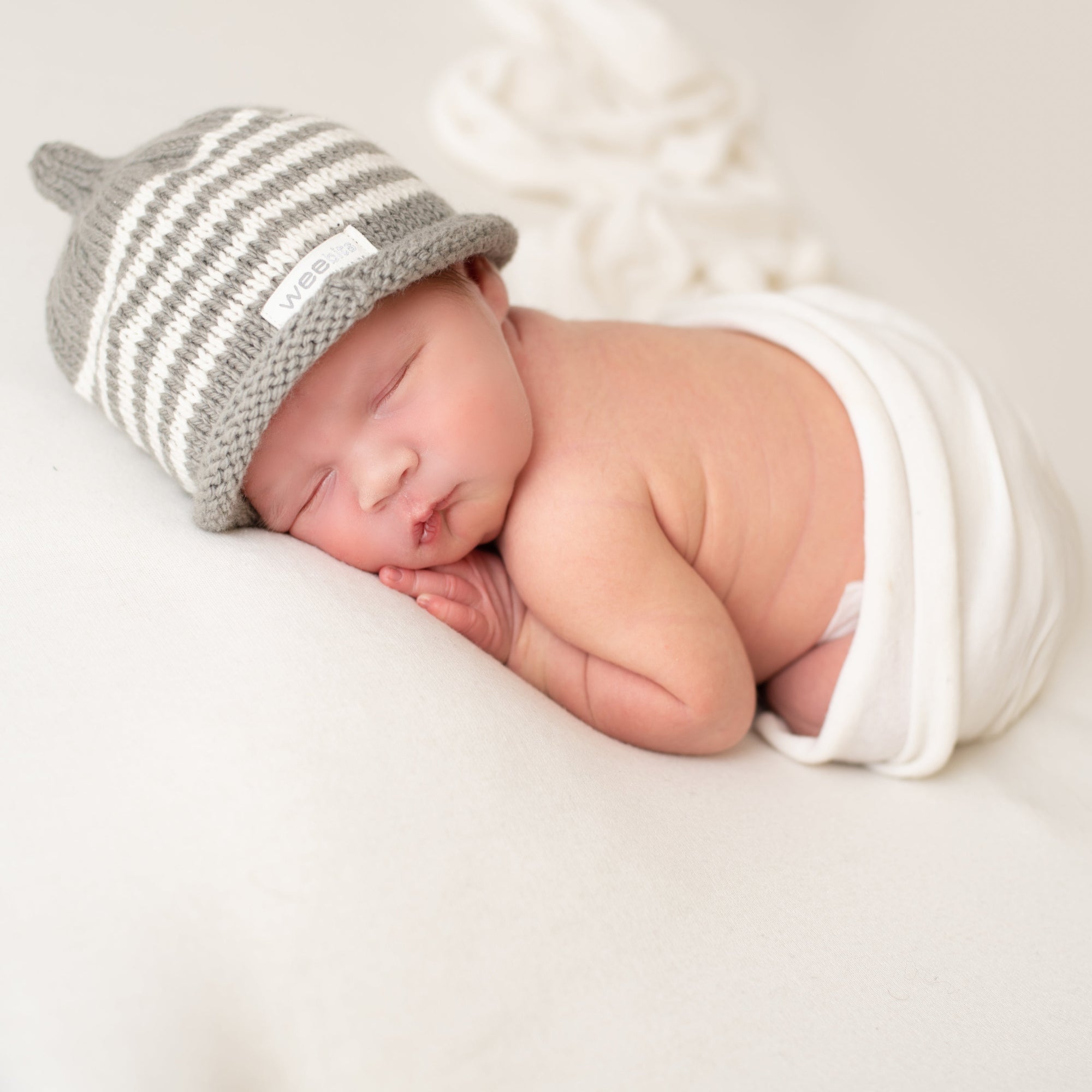 A sleeping newborn baby wrapped in a white swaddle, wearing a gray and white striped knit hat with a small white brand tag that reads 'Weebits'. The baby is lying on a soft white blanket.