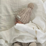 Newborn baby wrapped in a white blanket wearing a oatmeal cable knit hat with a pom-pom.