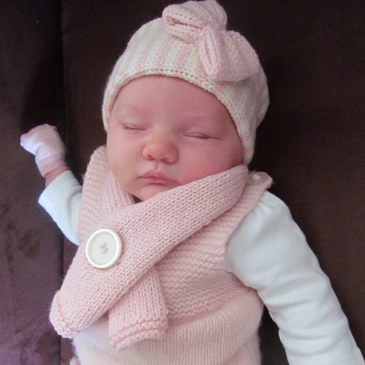 A sleeping newborn baby wearing a light pink headband hat with a bow, a matching knitted scarf with a large decorative button, and a pale pink sleeveless vest over a white long-sleeve shirt, lying on a dark brown surface.