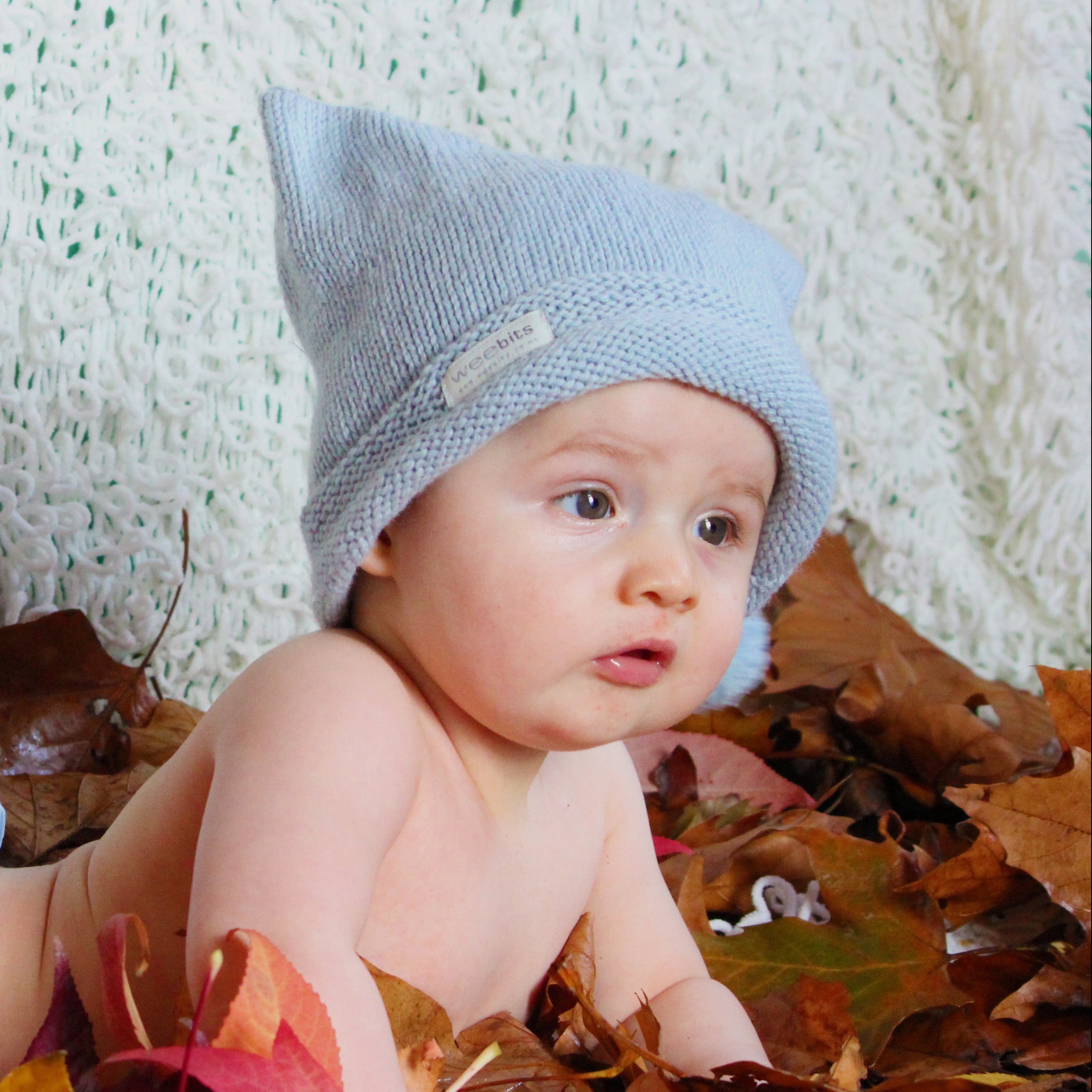 A baby lying on autumn leaves, wearing a light blue knit hat.