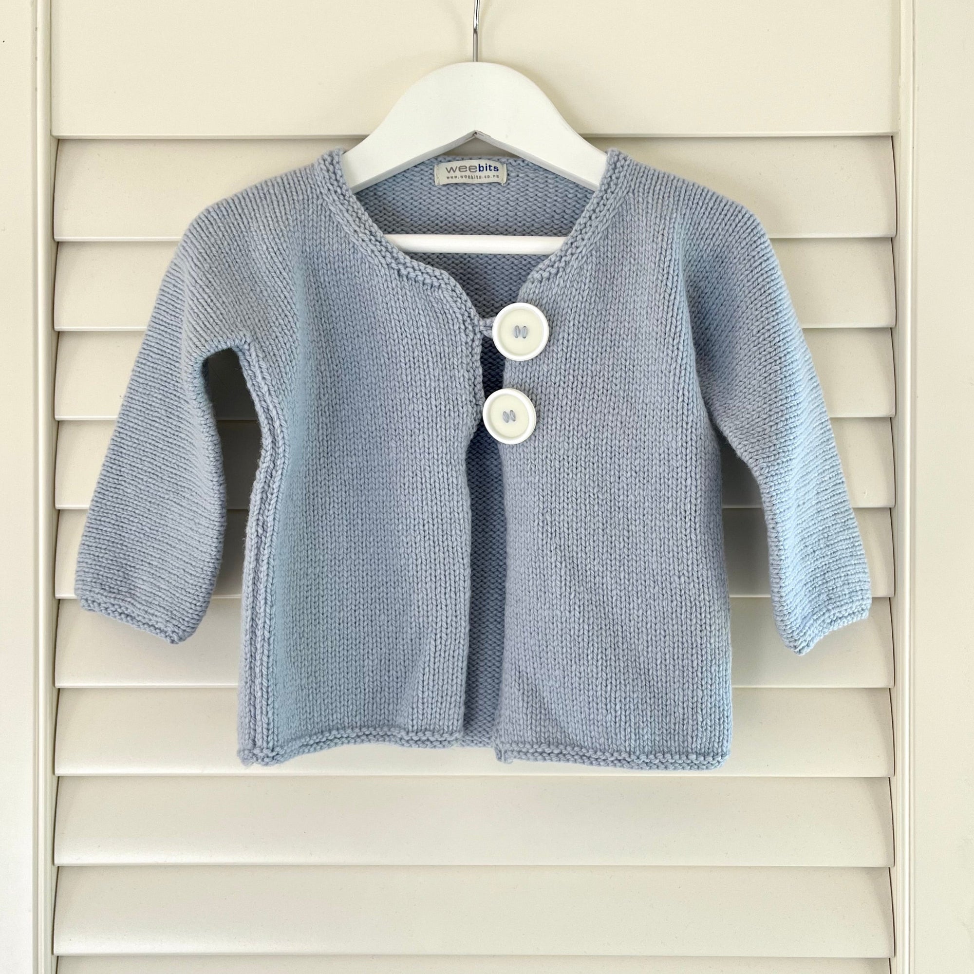 A blue knit baby cardigan sweater with two white buttons, hanging on a white hanger against a white wooden shutter background.