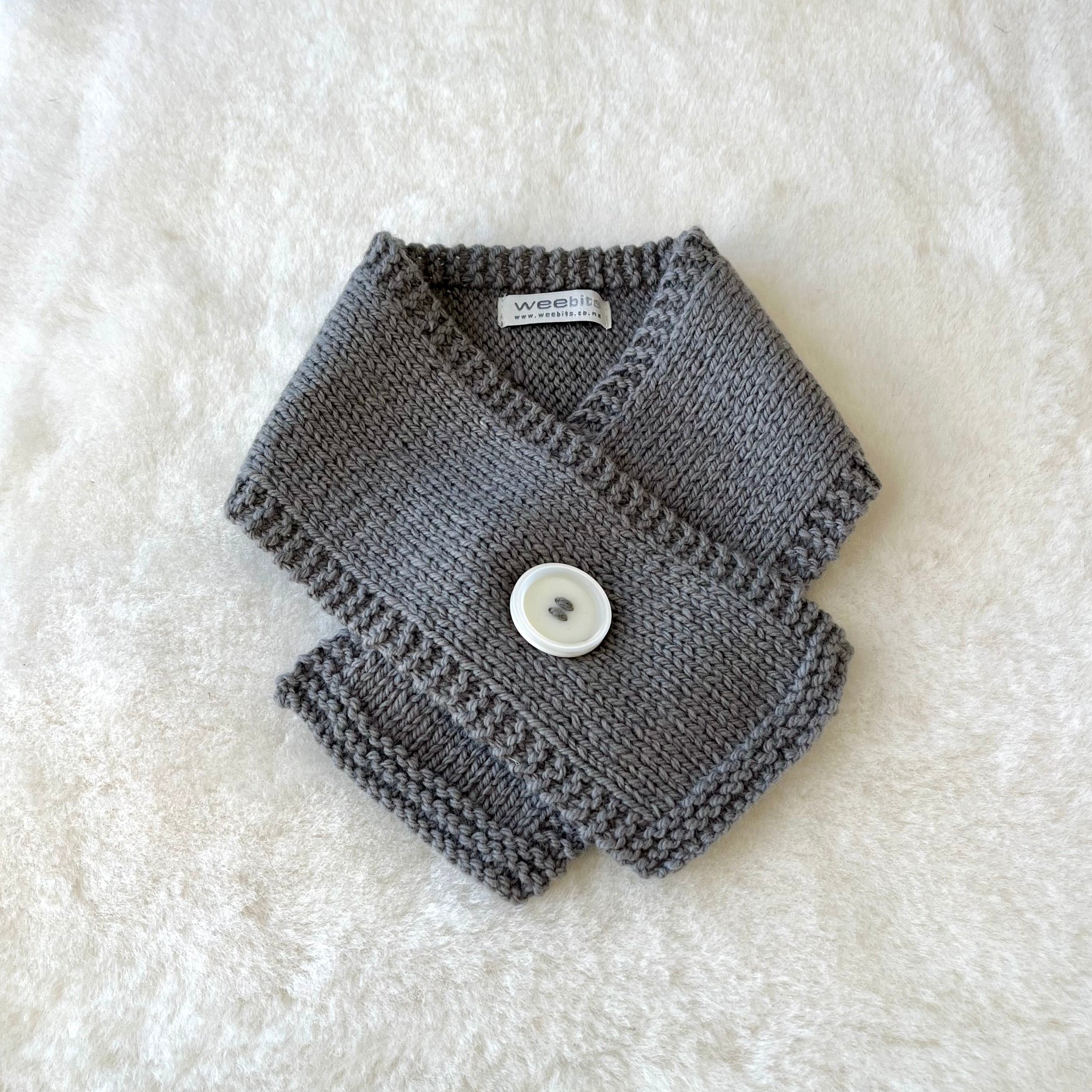 A mushroom knitted baby scarf with a white button closure, laid flat on a white textured faux fur background.