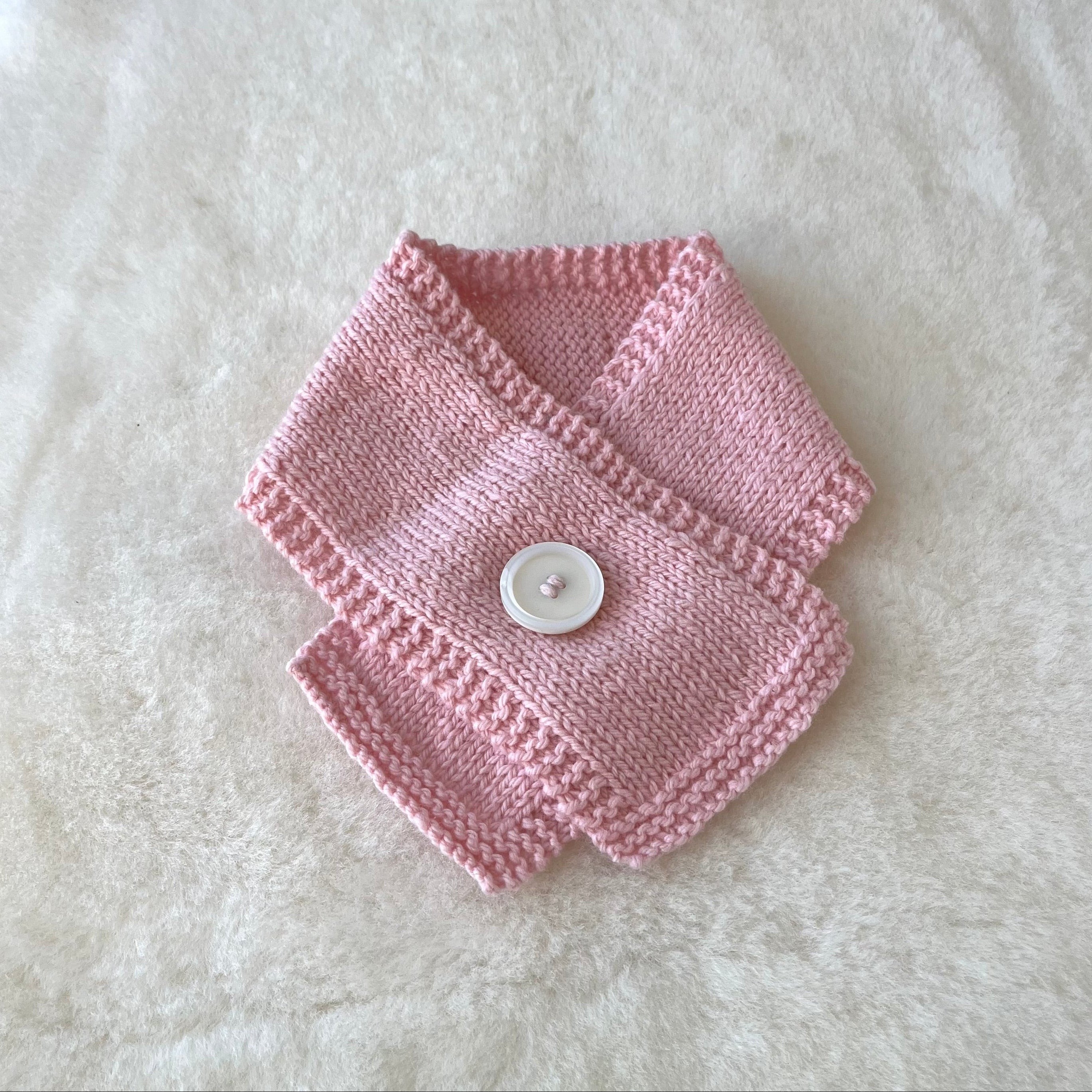 A pink knitted baby scarf with a white button closure, laid flat on a white textured faux fur background.