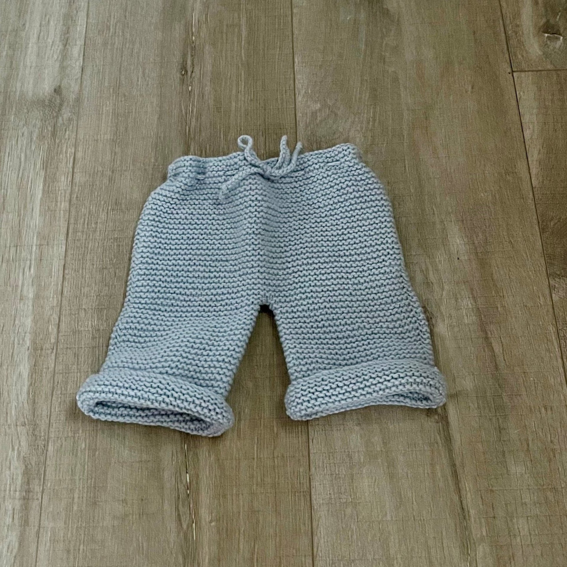 A pair of light blue hand-knit baby shorts with a matching drawstring waistband, laid flat on a white fabric background.