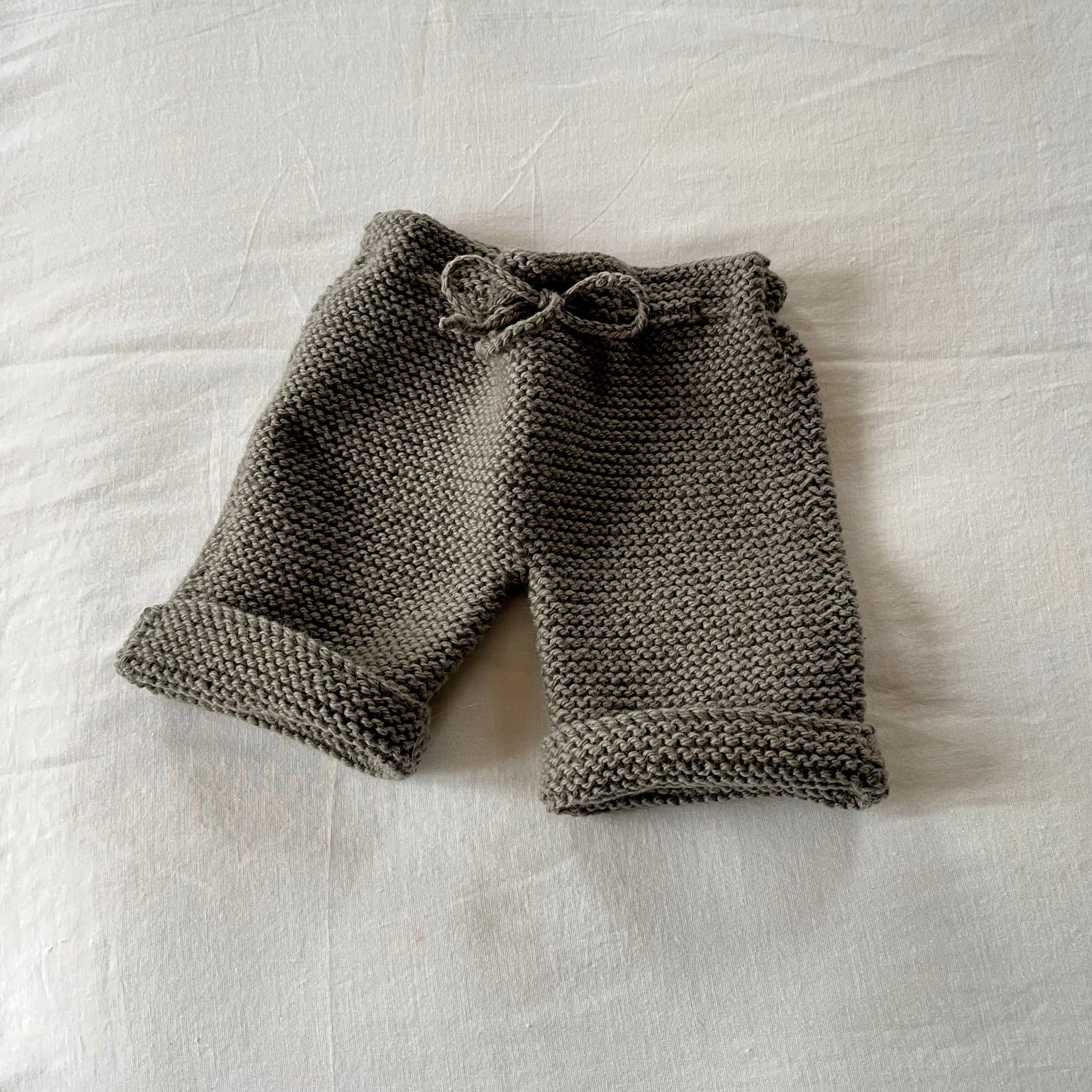 Hand-knit baby pants in a warm taupe-brown color, featuring a matching drawstring waistband and rolled cuffs, laid flat on a white fabric background.