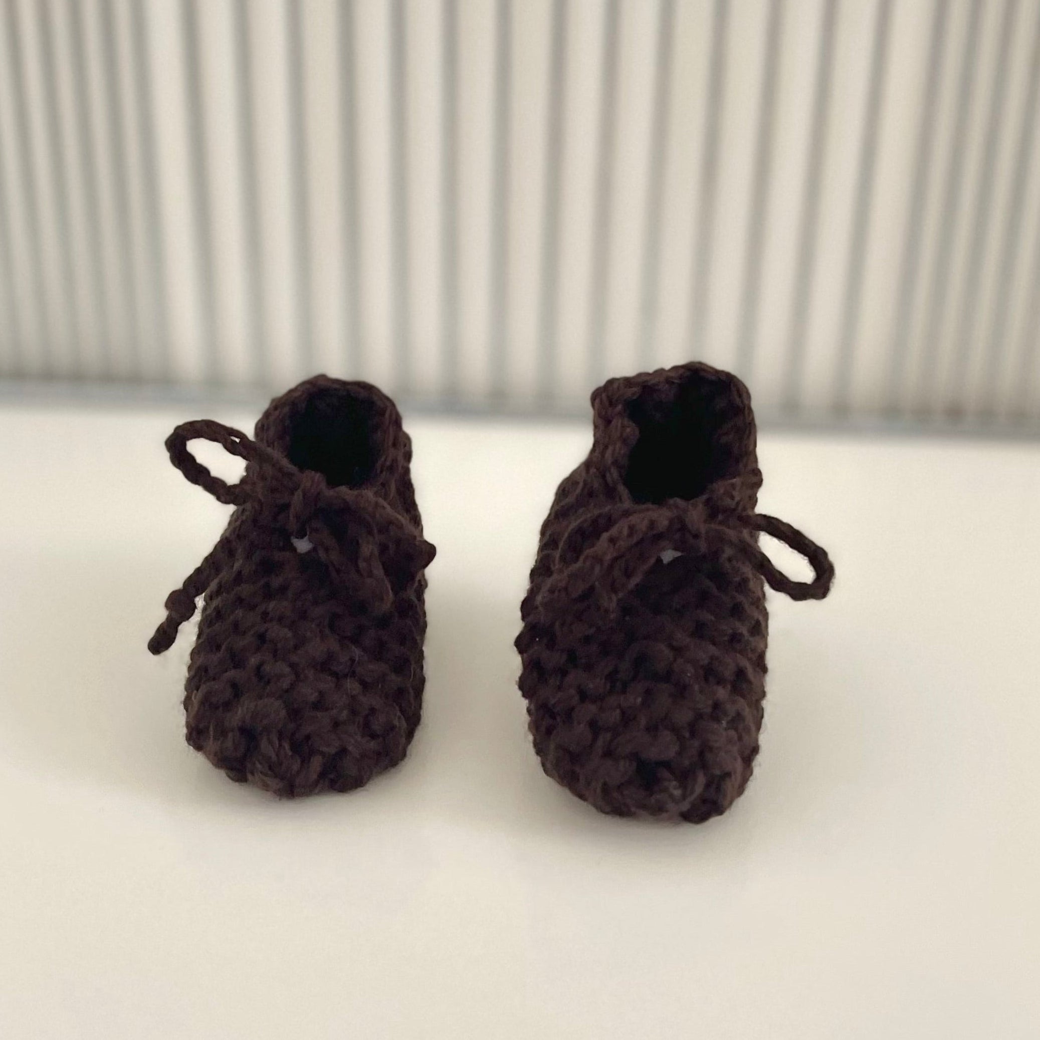 A pair of dark brown knitted baby booties with matching bows, displayed on a white surface in front of a silver ribbed background.