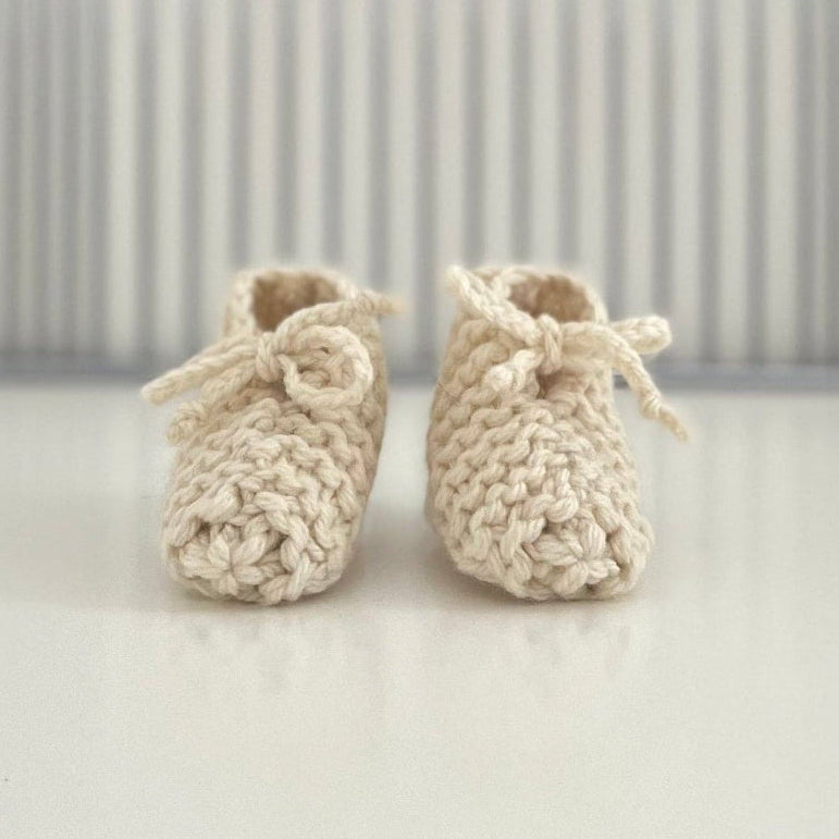 Pair of natural baby chunky knit booties on a white surface with a striped background