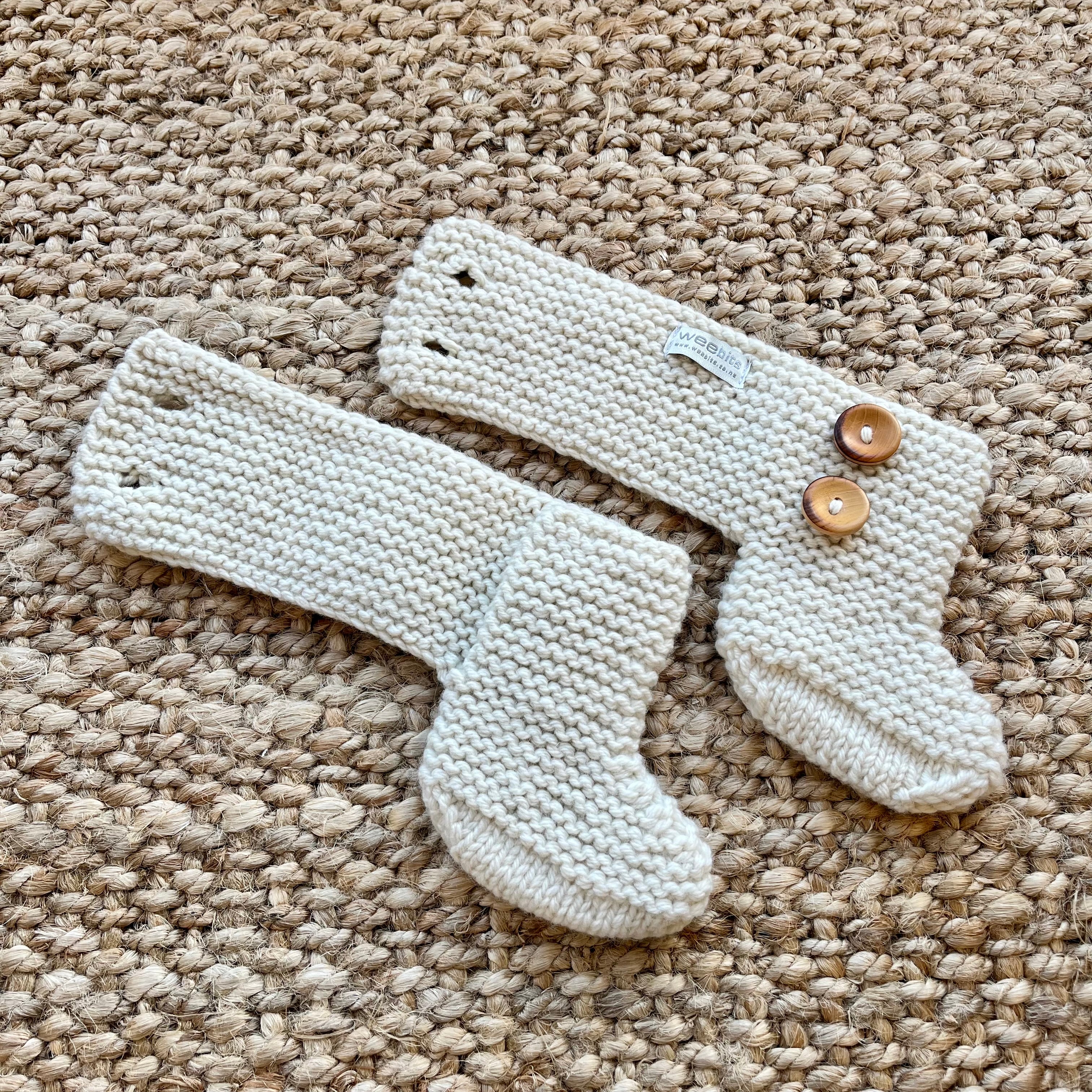 A pair of cream-colored hand-knit baby booties with two wooden buttons, laid flat on a textured beige woven rug.