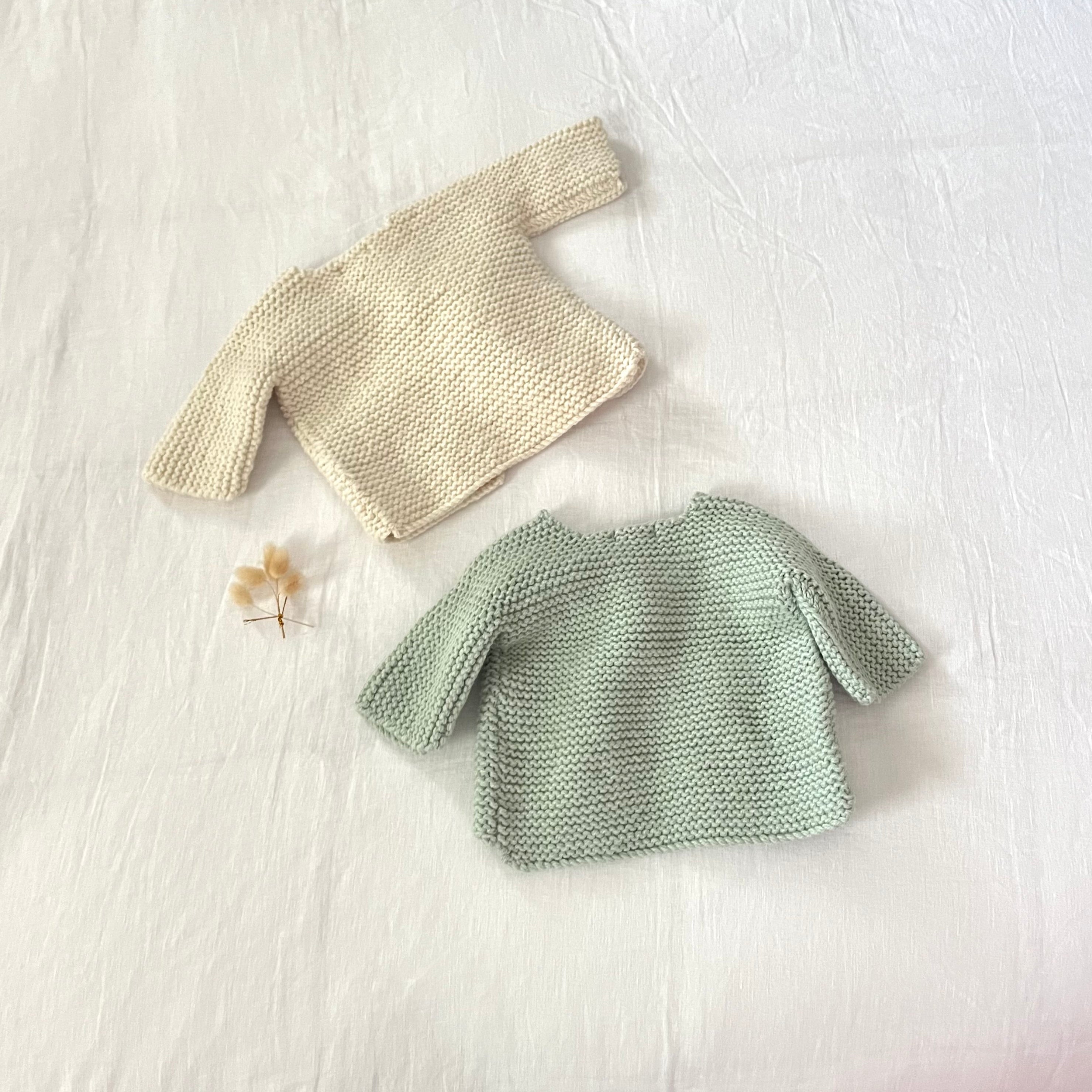 Two knitted baby cardigans, one natural and one green, on a light surface.
