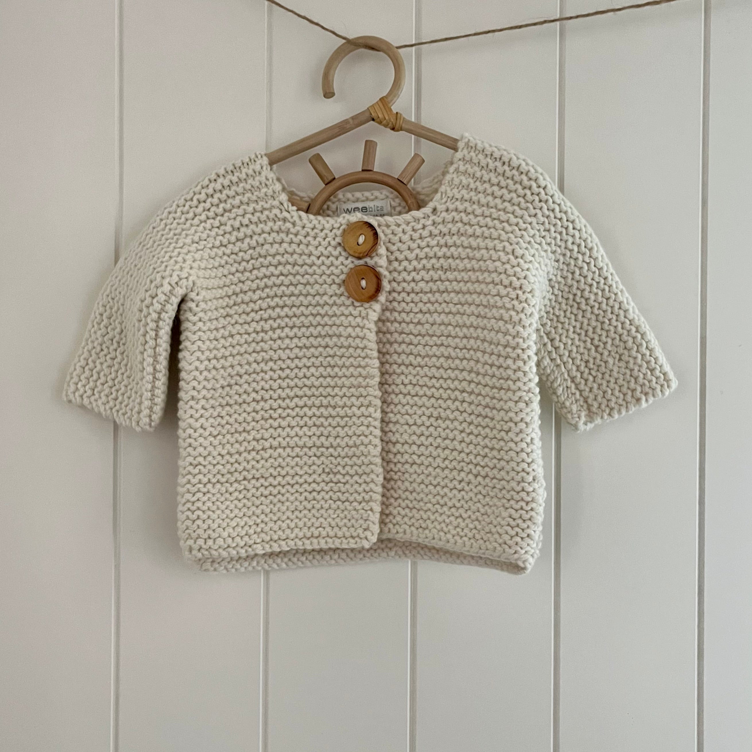 Knitted baby cardigan with wooden buttons on a hanger against a white paneled wall.