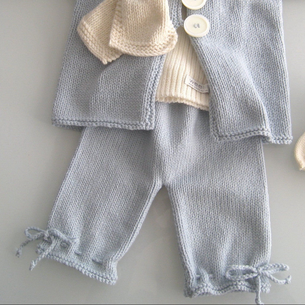 A baby knit clothing set laid flat on a light gray background, including a light blue-gray cardigan with cream buttons, matching knit pants, a cream knit scarf, a cream knit beret, and a pair of cream knit booties.
